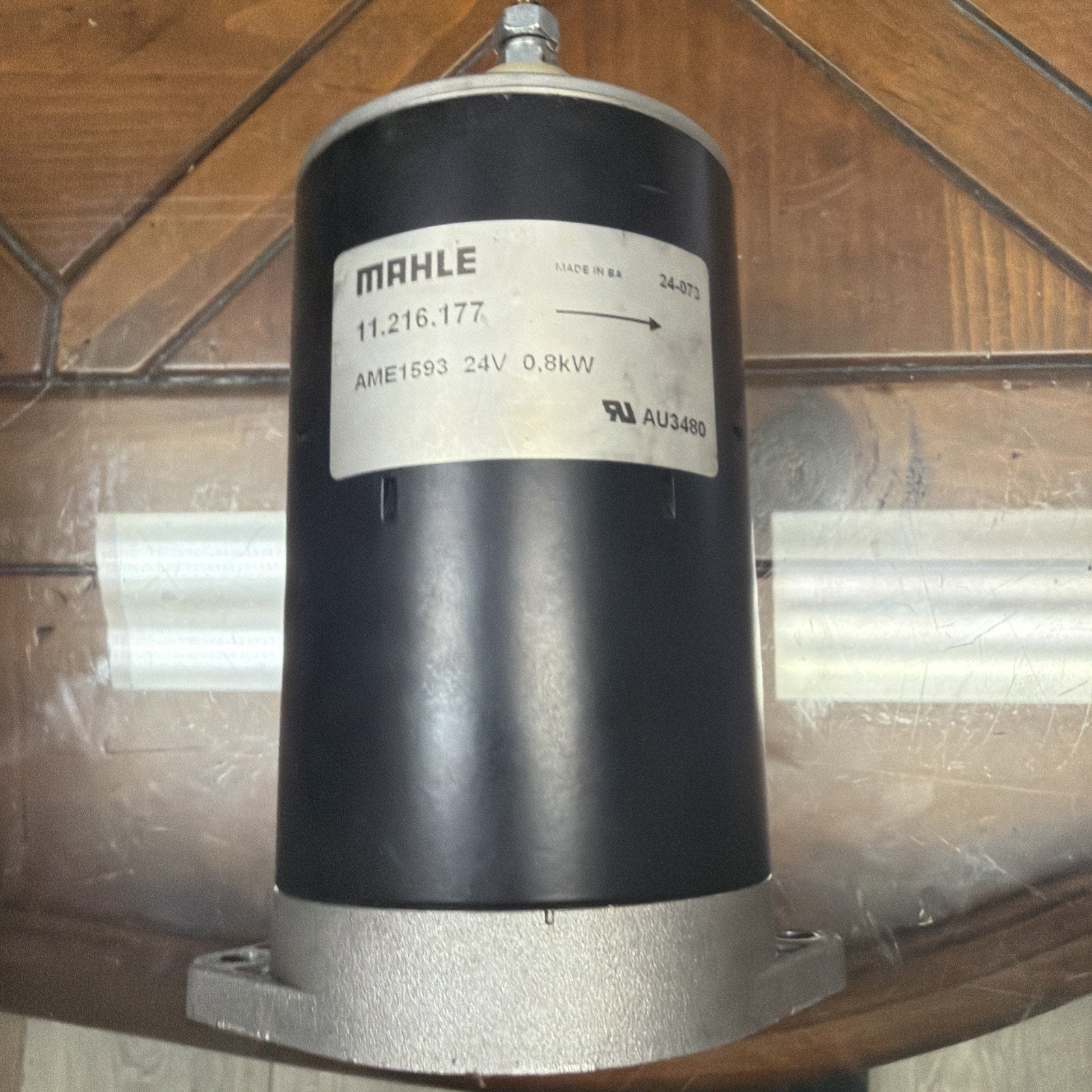 Cylindrical black and silver motor with a white label displaying "MAHLE," product code, voltage "24V," power rating "0.8kW," and certification marks, placed on a wooden and glass surface.