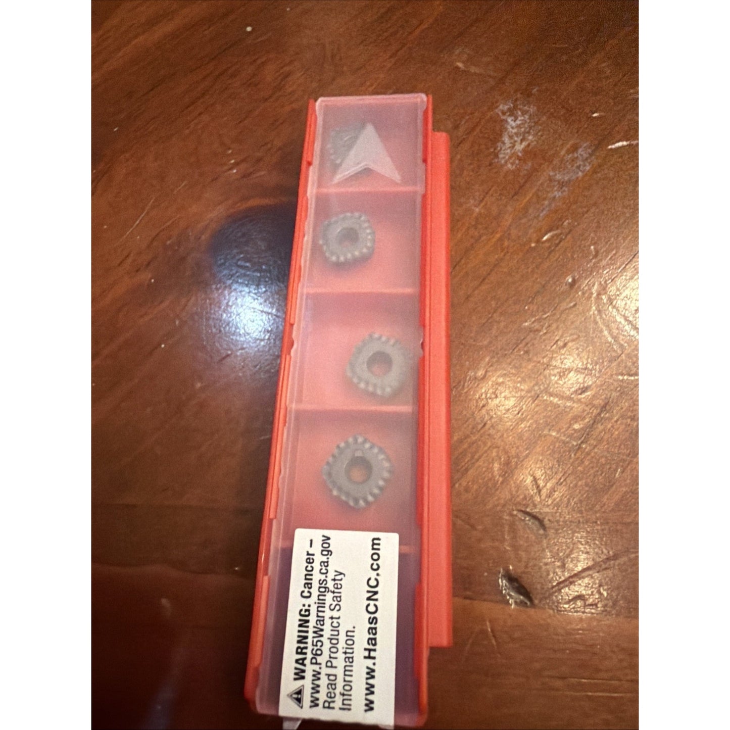 Narrow translucent red plastic container with four compartments, each holding a differently shaped metal cutting insert, placed on a wood surface with a safety warning label visible.