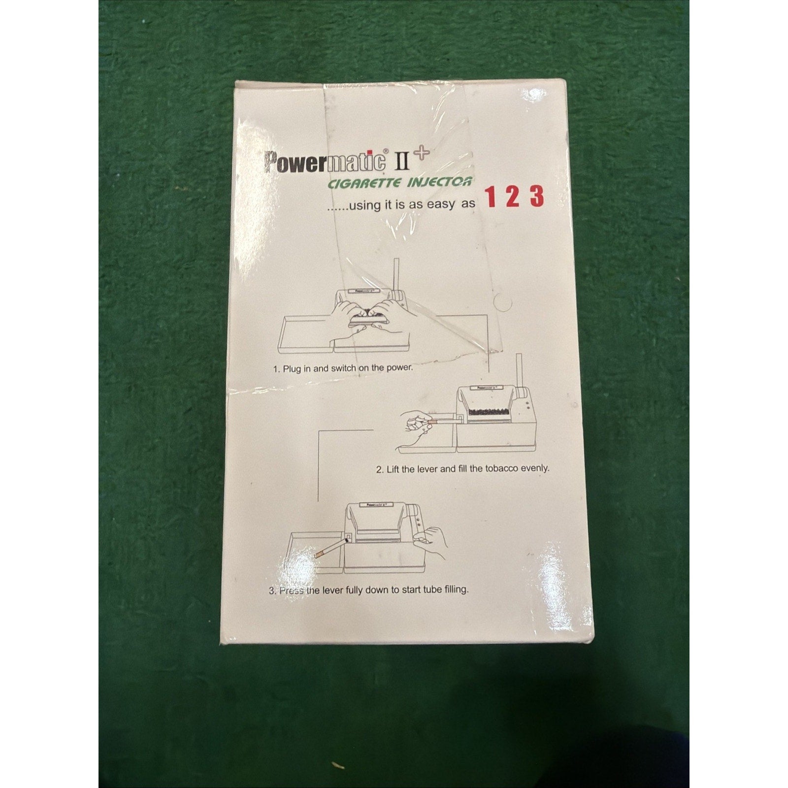 White product box featuring printed instructions and simple line drawings demonstrating the three-step process to use a Powermatic II+ cigarette injector machine.