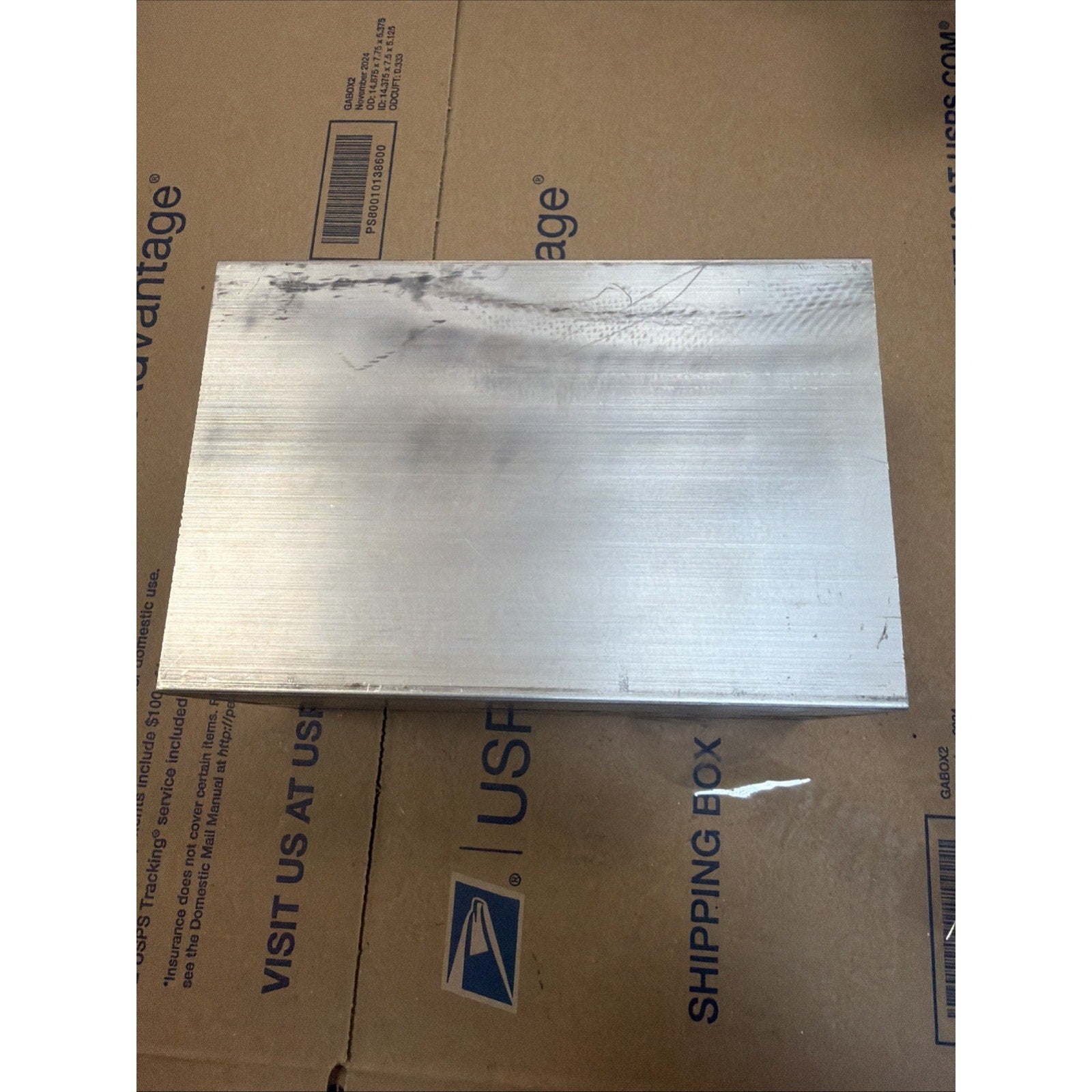 Rectangular metallic block with a smooth surface showing faint horizontal machining marks and minor discoloration at the top left corner, placed on a USPS shipping box.