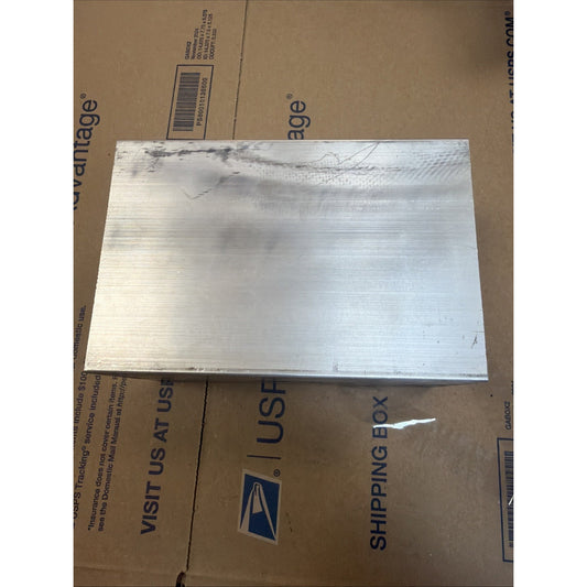 Rectangular metallic block with a smooth surface showing faint horizontal machining marks and minor discoloration at the top left corner, placed on a USPS shipping box.
