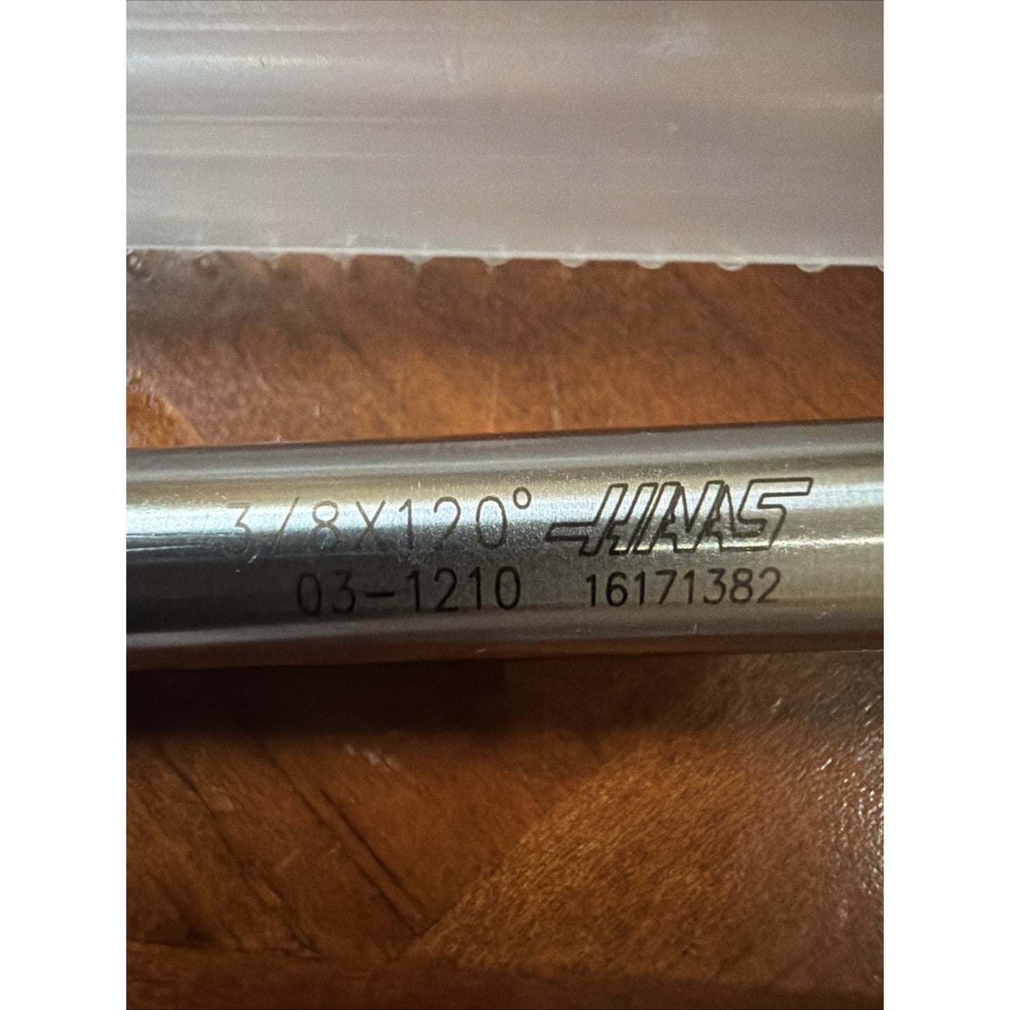 Close-up of a metal cylindrical tool with engraved markings including "3/8 x 120°," a product code, and the Haas logo, resting on a wooden surface.