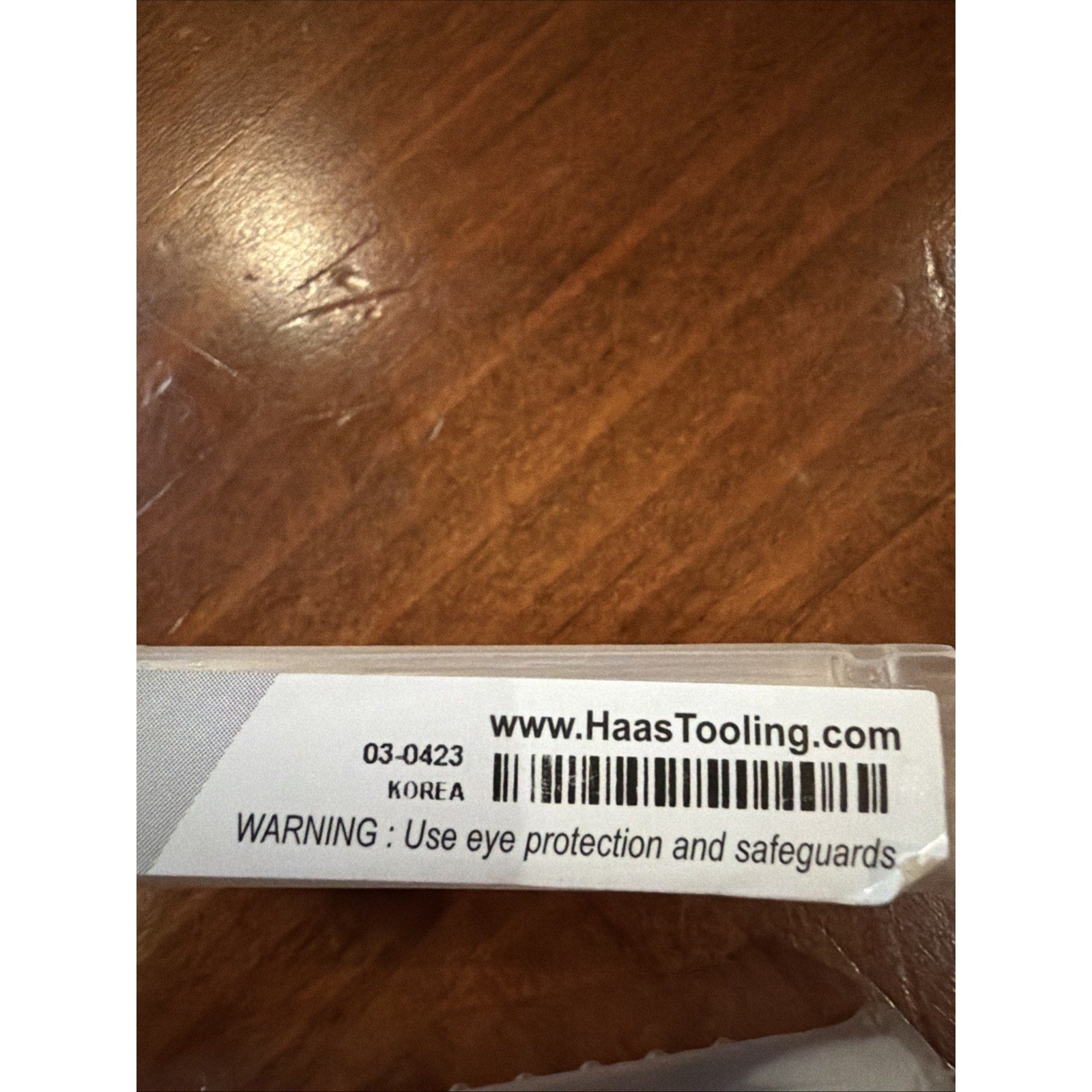 Close-up of a white label on a clear plastic container showing barcode, "www.HaasTooling.com," product code "03-0423," origin marked "KOREA," and a warning to use eye protection and safeguards, against a wooden surface.