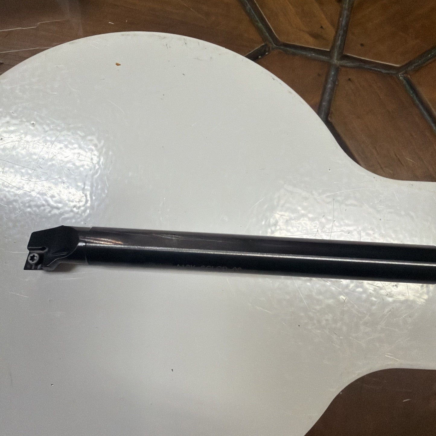 Long black metal cutting tool with an angled tip and a small screw, resting diagonally on a glossy white surface with a brown tiled floor partially visible in the background.