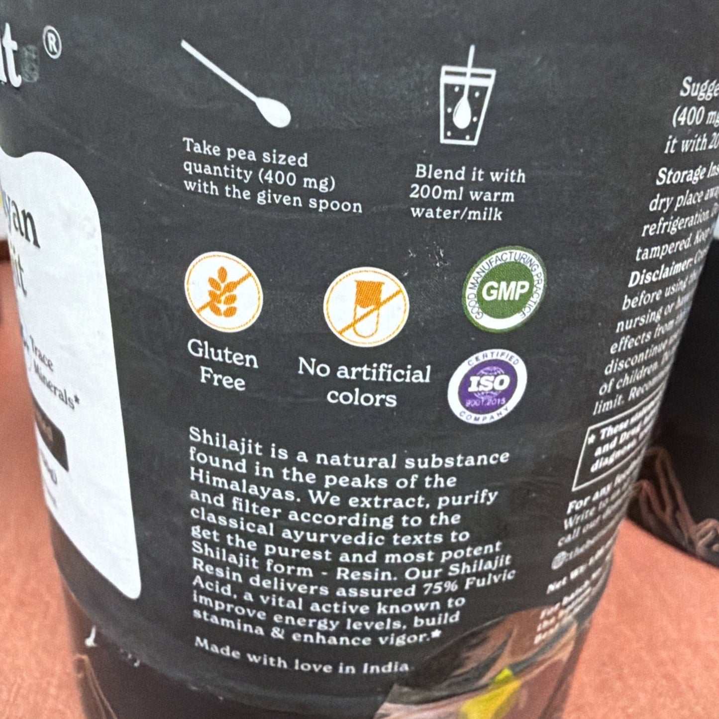 Close-up of a black product label showing instructions to take 400 mg with a spoon and blend with 200 ml warm water or milk, along with icons for gluten-free, no artificial colors, GMP and ISO certification, and text about Shilajit resin benefits.