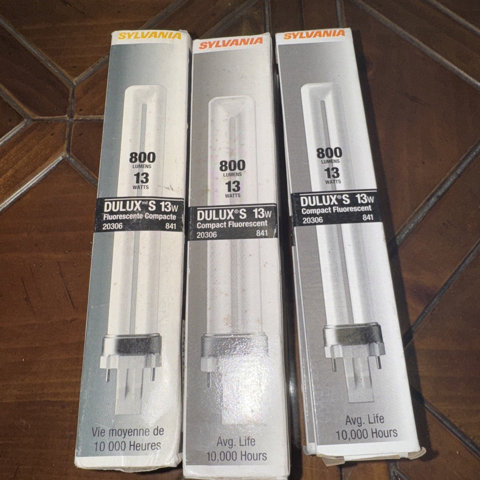 Three Sylvania DULUX S 13 watt 800 lumen compact fluorescent bulbs in original packaging on wooden surface