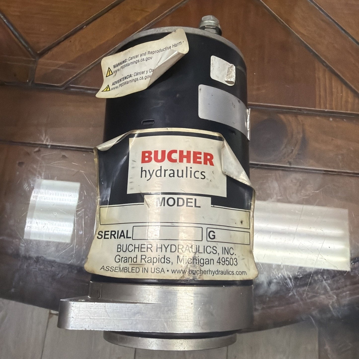 Black cylindrical hydraulic component with peeling labels, including a prominent "BUCHER hydraulics" logo, serial number section, and warning stickers, resting on a glass surface.