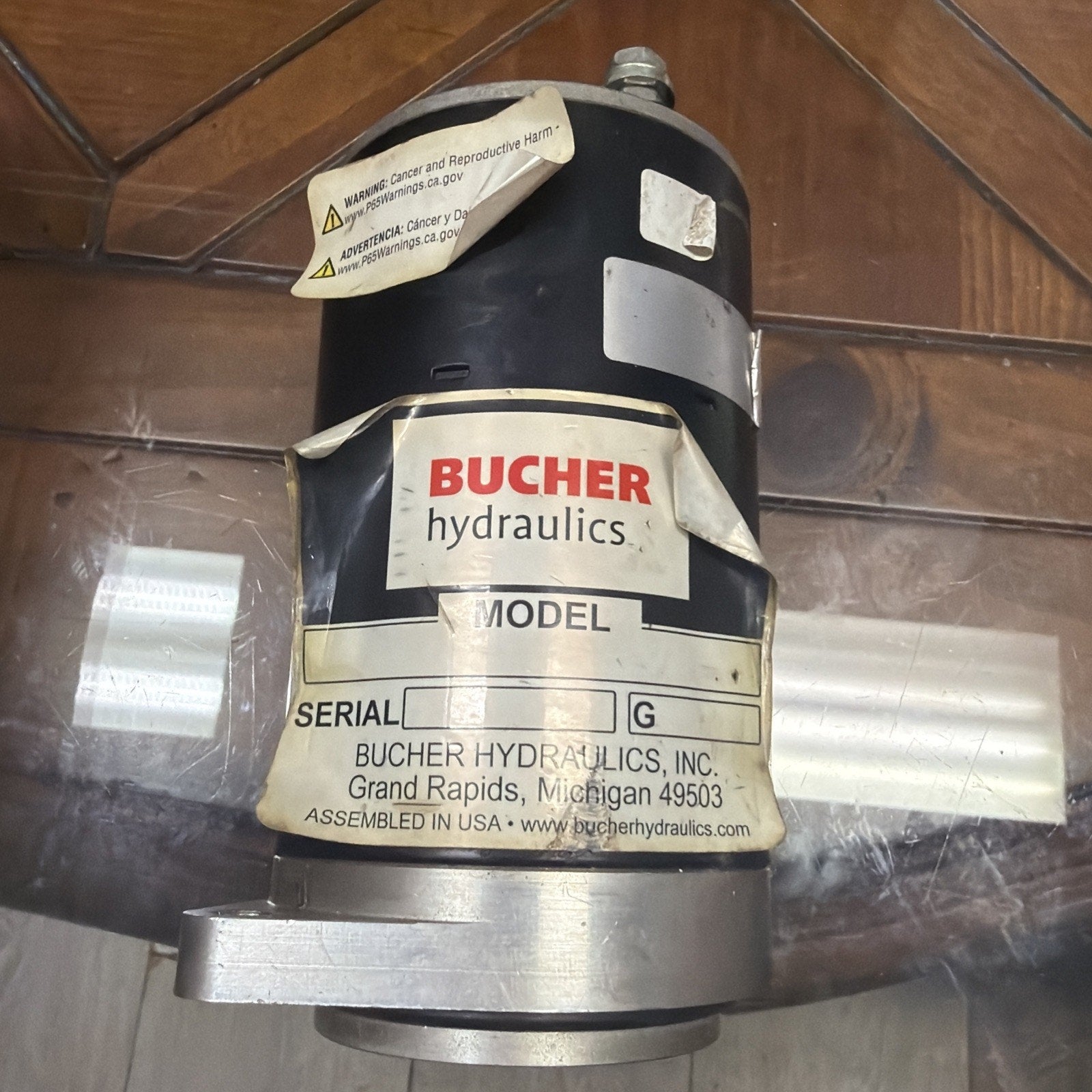 Black cylindrical hydraulic component with peeling labels, including a prominent "BUCHER hydraulics" logo, serial number section, and warning stickers, resting on a glass surface.