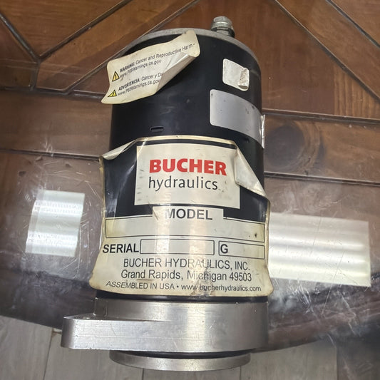 Black cylindrical hydraulic component with peeling labels, including a prominent "BUCHER hydraulics" logo, serial number section, and warning stickers, resting on a glass surface.