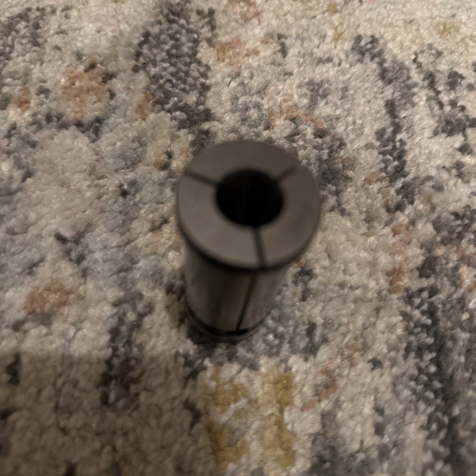 Close-up of a small metal cylindrical collet with three vertical slits, resting upright on a textured multicolor carpet surface.
