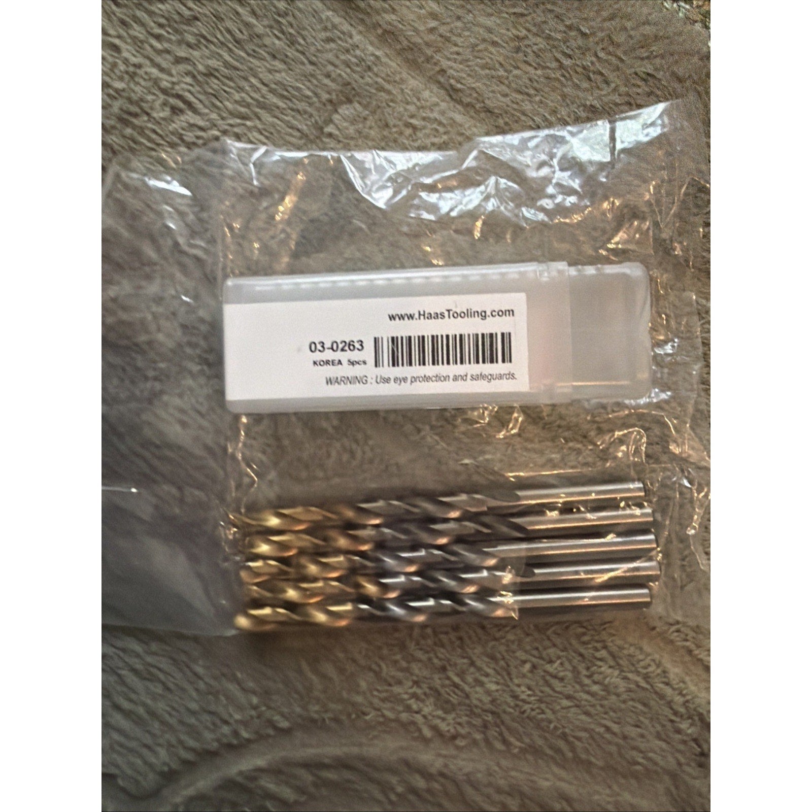 Five shiny metal drill bits with spiral grooves, resting on a clear plastic package labeled with a barcode and a warning notice, placed on a textured fabric surface.