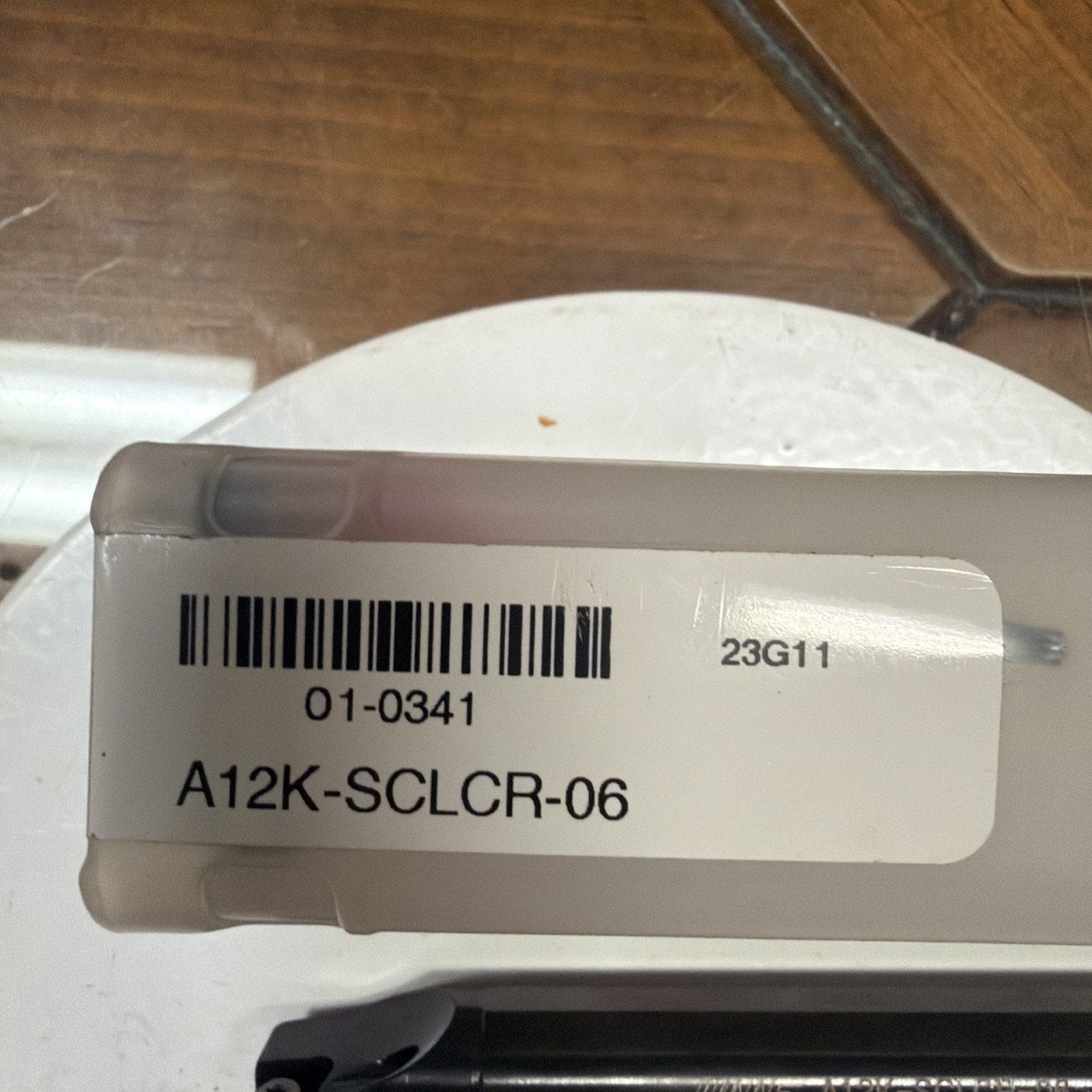 White rectangular label with black printed barcode and alphanumeric code "A12K-SCLCR-06" and "01-0341" on a semi-transparent plastic container placed on a white circular surface over a wooden table.