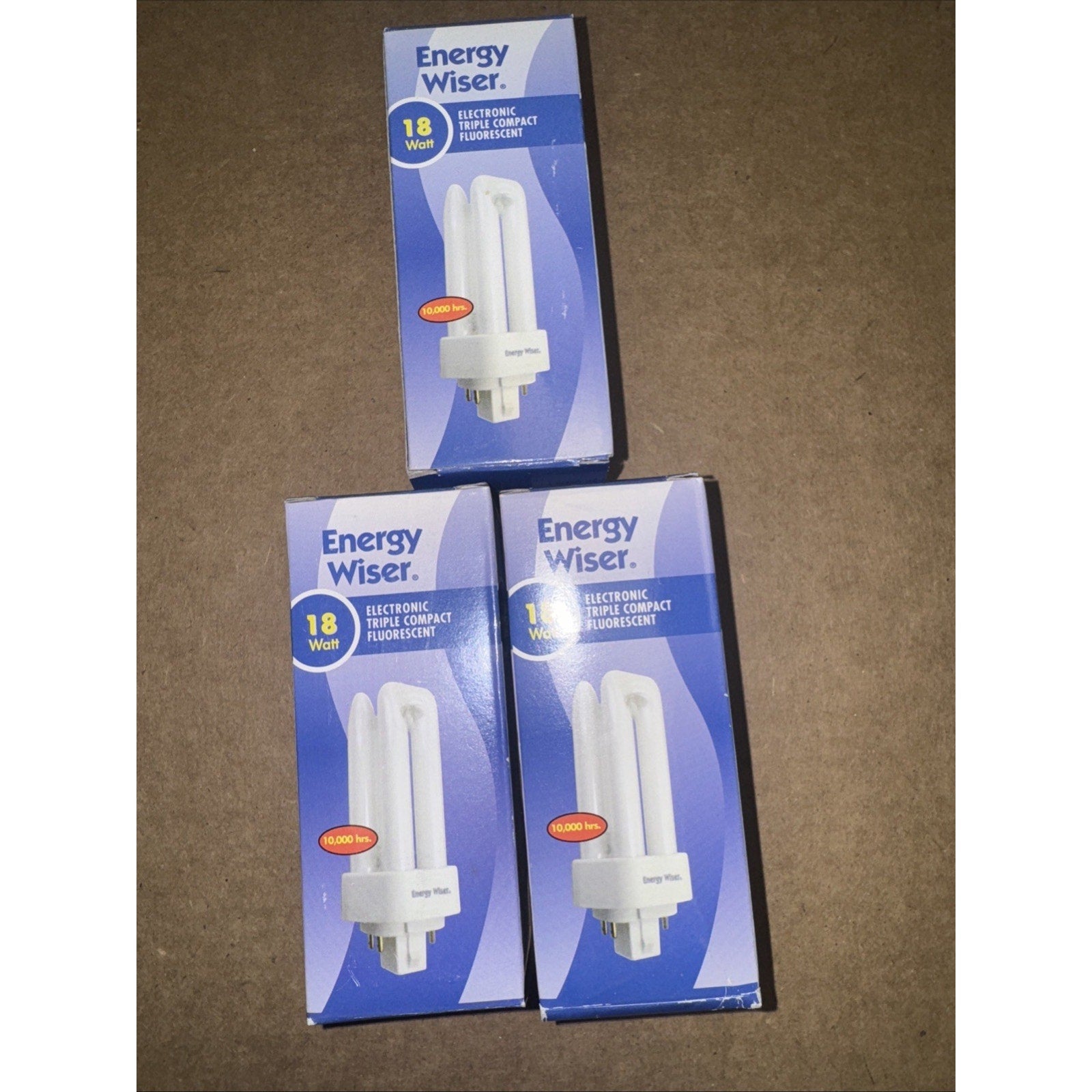 Three Energy Wiser 18W electronic triple compact fluorescent replacement bulbs in blue and white packaging on brown surface