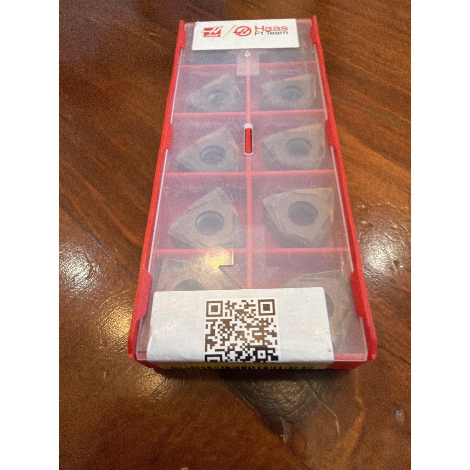 Red plastic case containing eight triangular metal cutting inserts with a translucent cover and labels featuring Haas and a QR code, placed on a wooden surface.