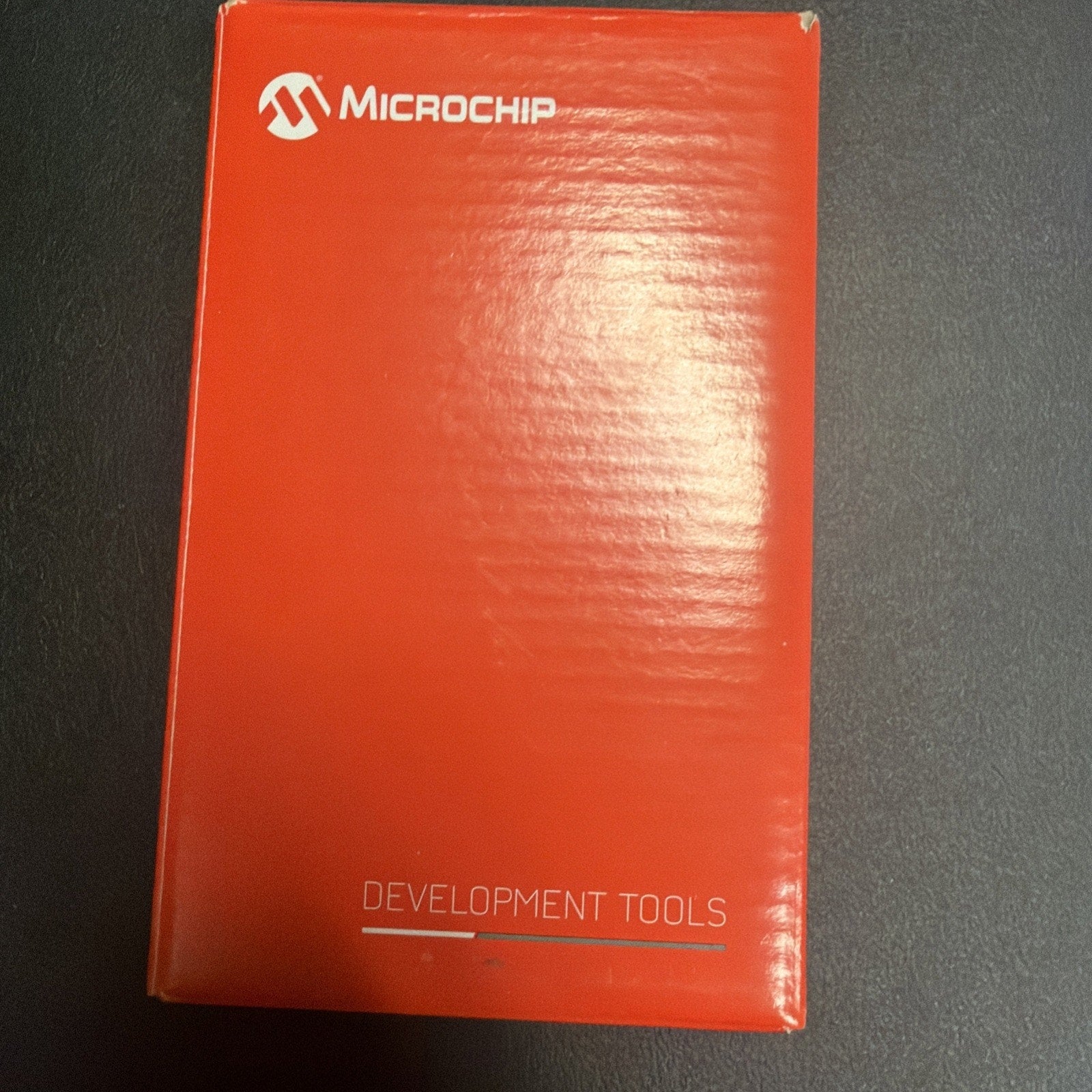 Red rectangular Microchip product box with a white logo and the words "Development Tools" printed on the front, placed on a dark textured surface.