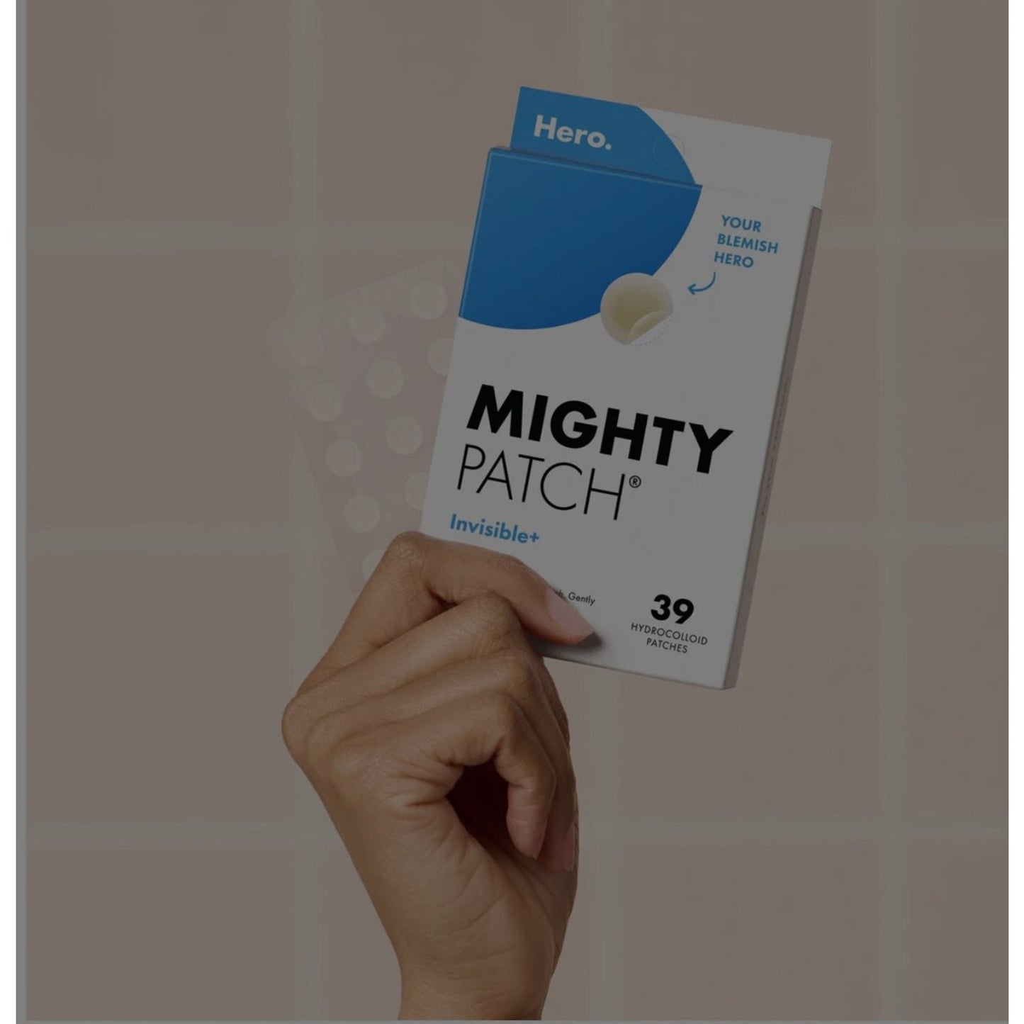 Hand holding a white and blue rectangular box labeled Mighty Patch Invisible+ featuring a single hydrocolloid patch on the front and text indicating 39 patches included.