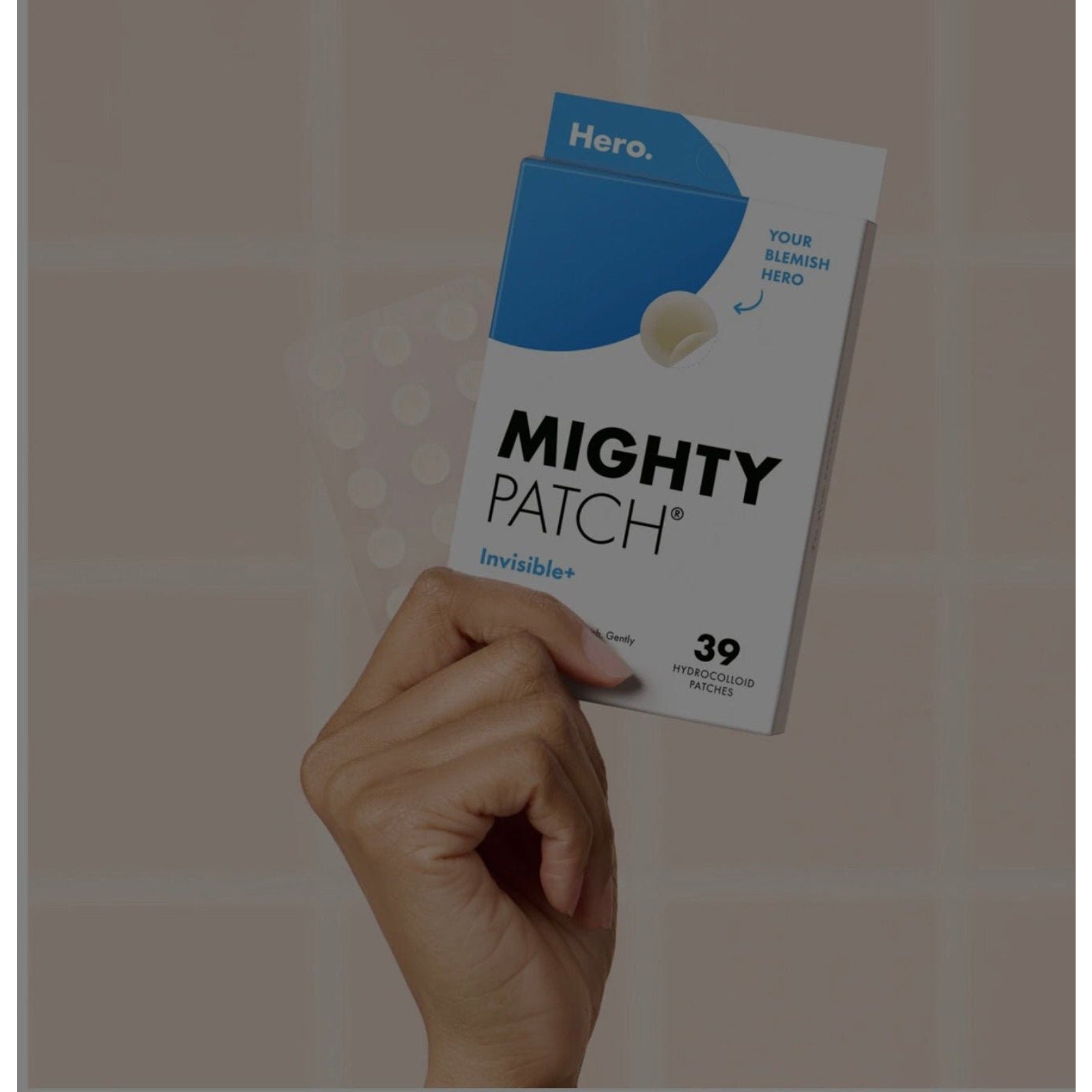 Hand holding a white and blue rectangular box labeled Mighty Patch Invisible+ featuring a single hydrocolloid patch on the front and text indicating 39 patches included.