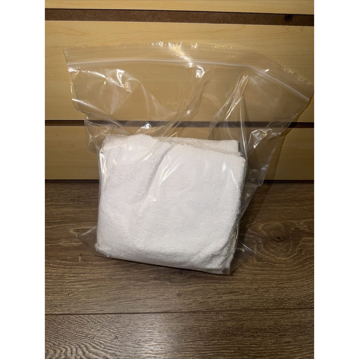 White terry cloth folded inside a clear resealable plastic bag placed on a wooden surface with light wood paneling in the background.