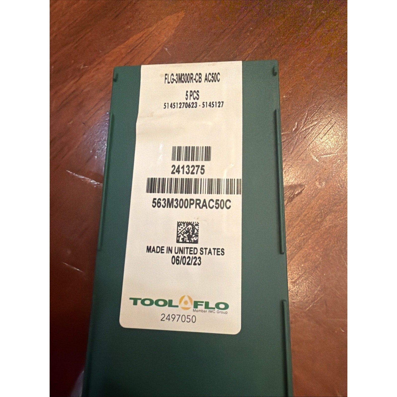 Dark green rectangular container with a white label displaying product code "FLGAK30DR-CB AC30C," quantity "5 PCS," barcodes, manufacturing date "06/02/23," and "Made in United States," placed on a wooden surface.