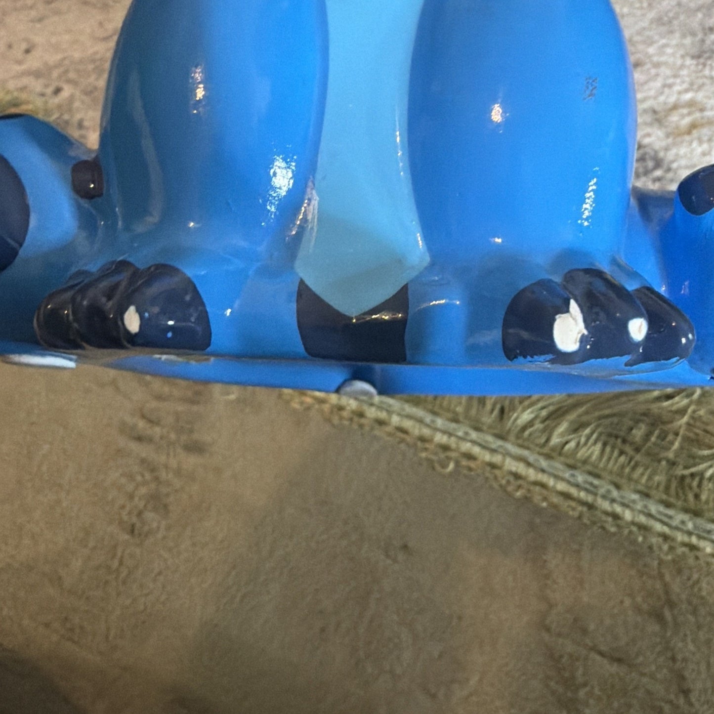 Close-up of a blue plastic figure showing detailed black and white painted feet and part of the body, resting on a textured beige carpet with a woven rug edge visible.