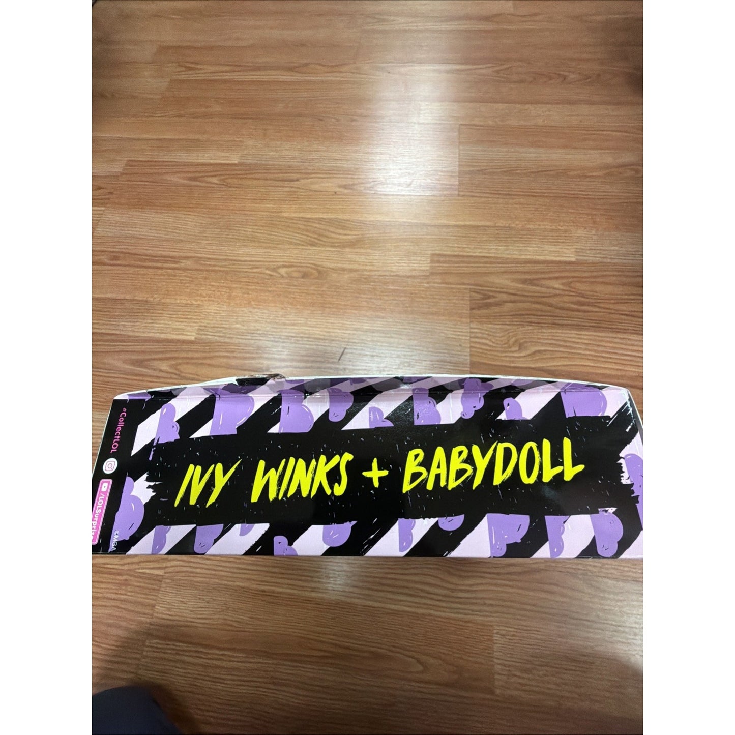 Rectangular product box with a bold purple and black diagonal striped design, featuring bright yellow text that reads "Ivy Winks + Babydoll," placed on a wooden floor.
