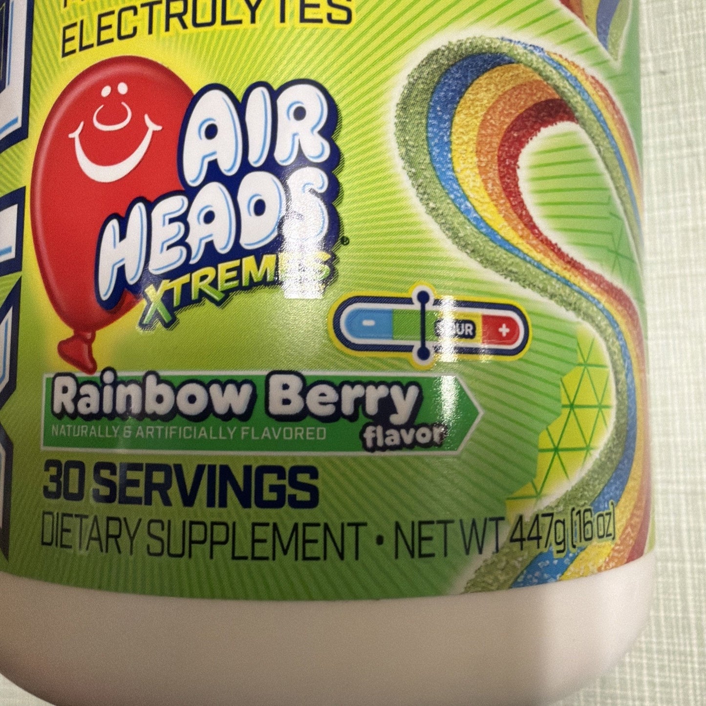 Close-up of a dietary supplement label for Air Heads Xtremes Rainbow Berry flavor highlighting 30 servings, natural and artificial flavoring, and electrolyte content on a green background with a colorful rainbow swirl.