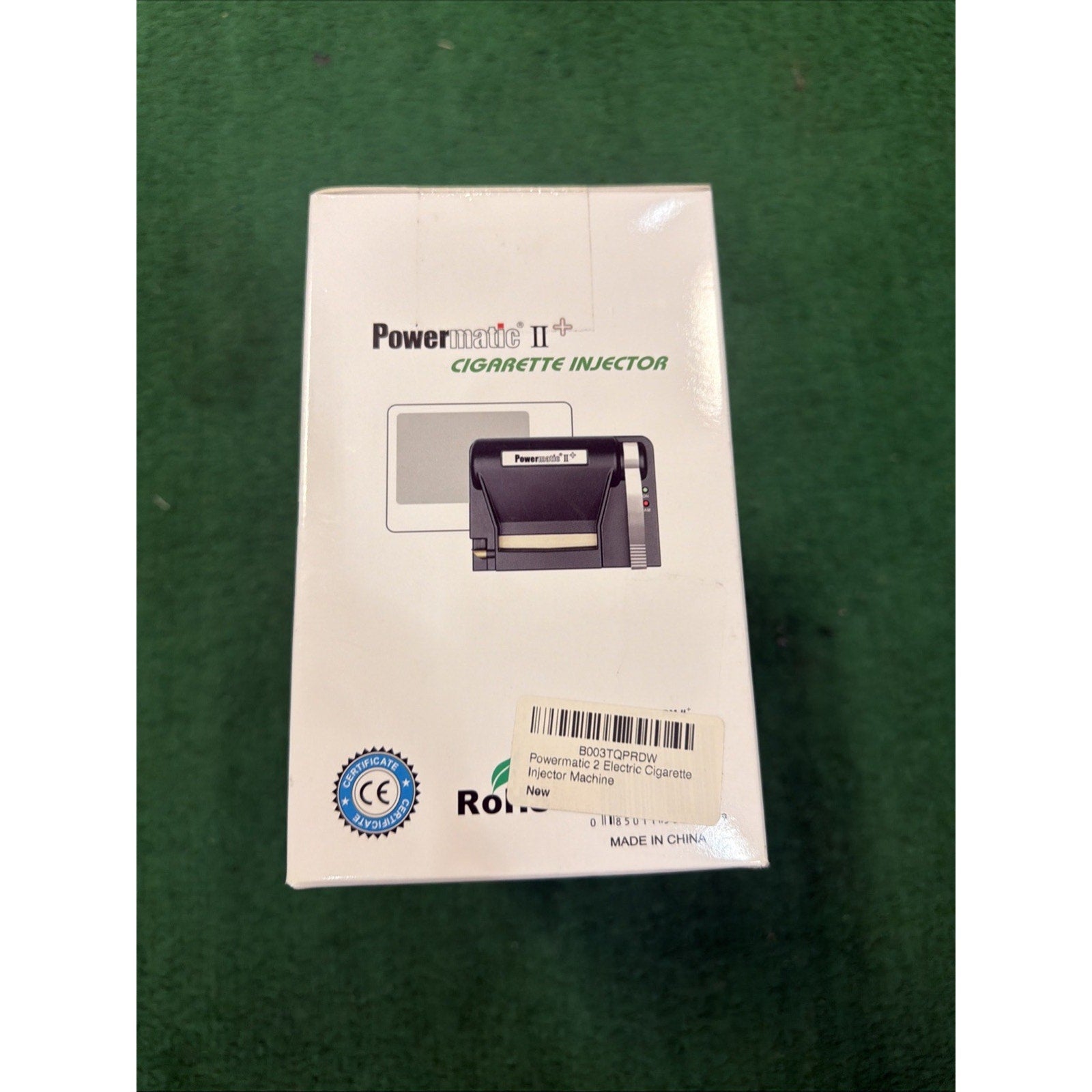 White rectangular box packaging for a Powermatic II+ cigarette injector machine featuring a small product image, certification mark, and a barcode sticker on a green surface.