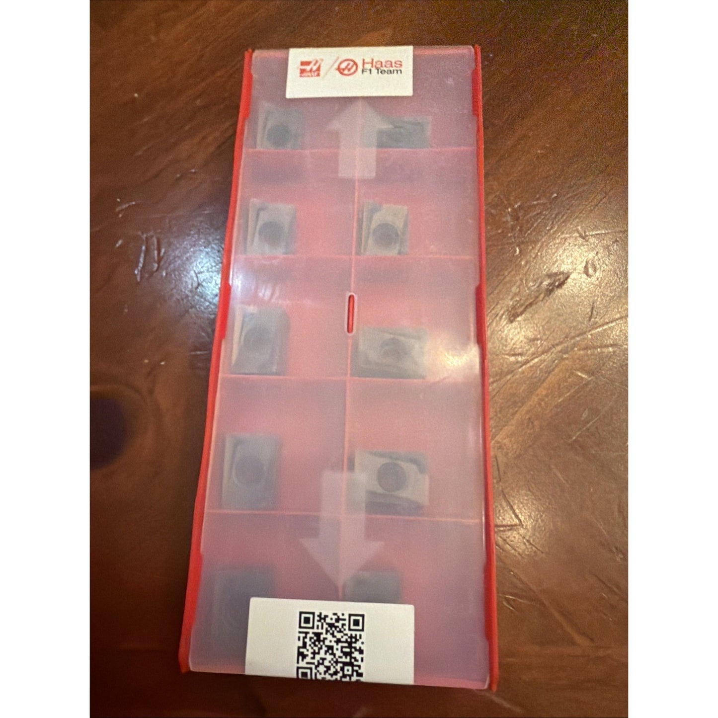 Tall translucent red plastic storage box with multiple compartments holding small metal inserts, featuring a Haas H1 Team label and a QR code, placed on a wooden surface.