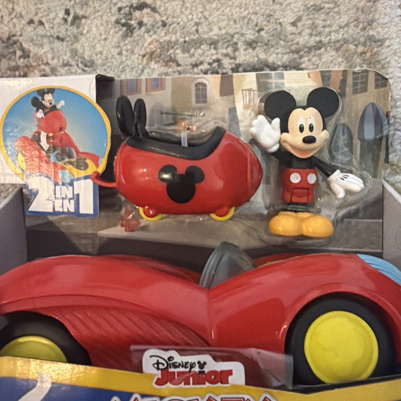 Red toy car with yellow wheels and a separate figurine of Mickey Mouse wearing red shorts and yellow shoes, displayed in packaging against a cityscape background.