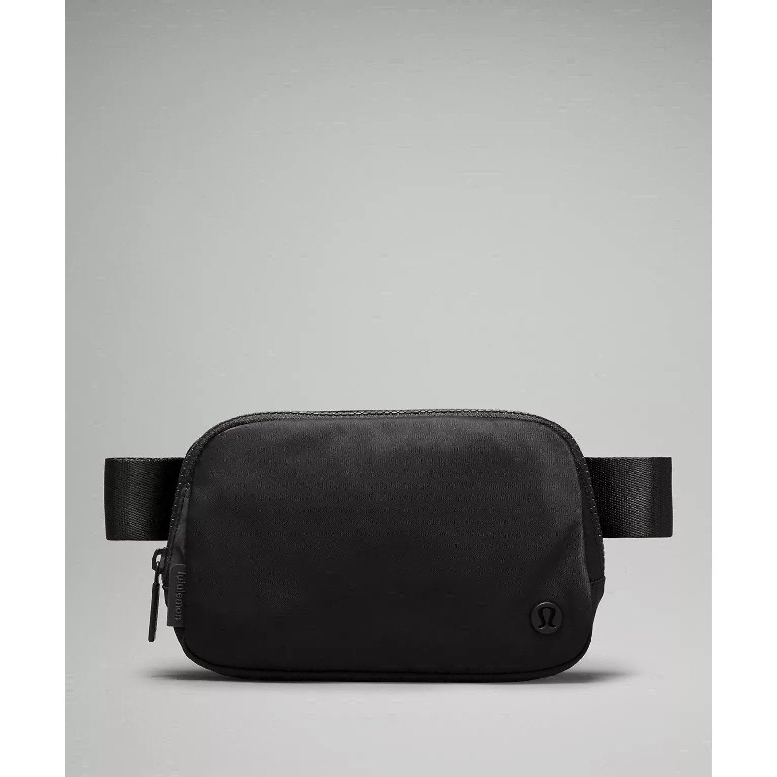 Minimalist black waist bag with a smooth matte finish, rounded rectangular shape, front zipper closure, and a small embossed logo on the bottom right corner.