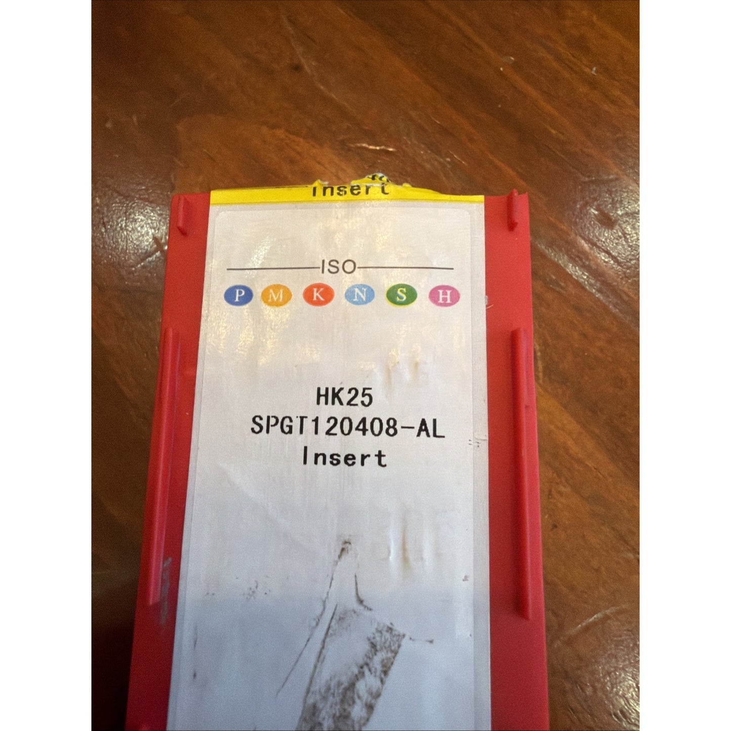 Red plastic storage container with a white label showing ISO color-coded classifications and text "HK25 SPGT120408-AL Insert," placed on a wooden surface.