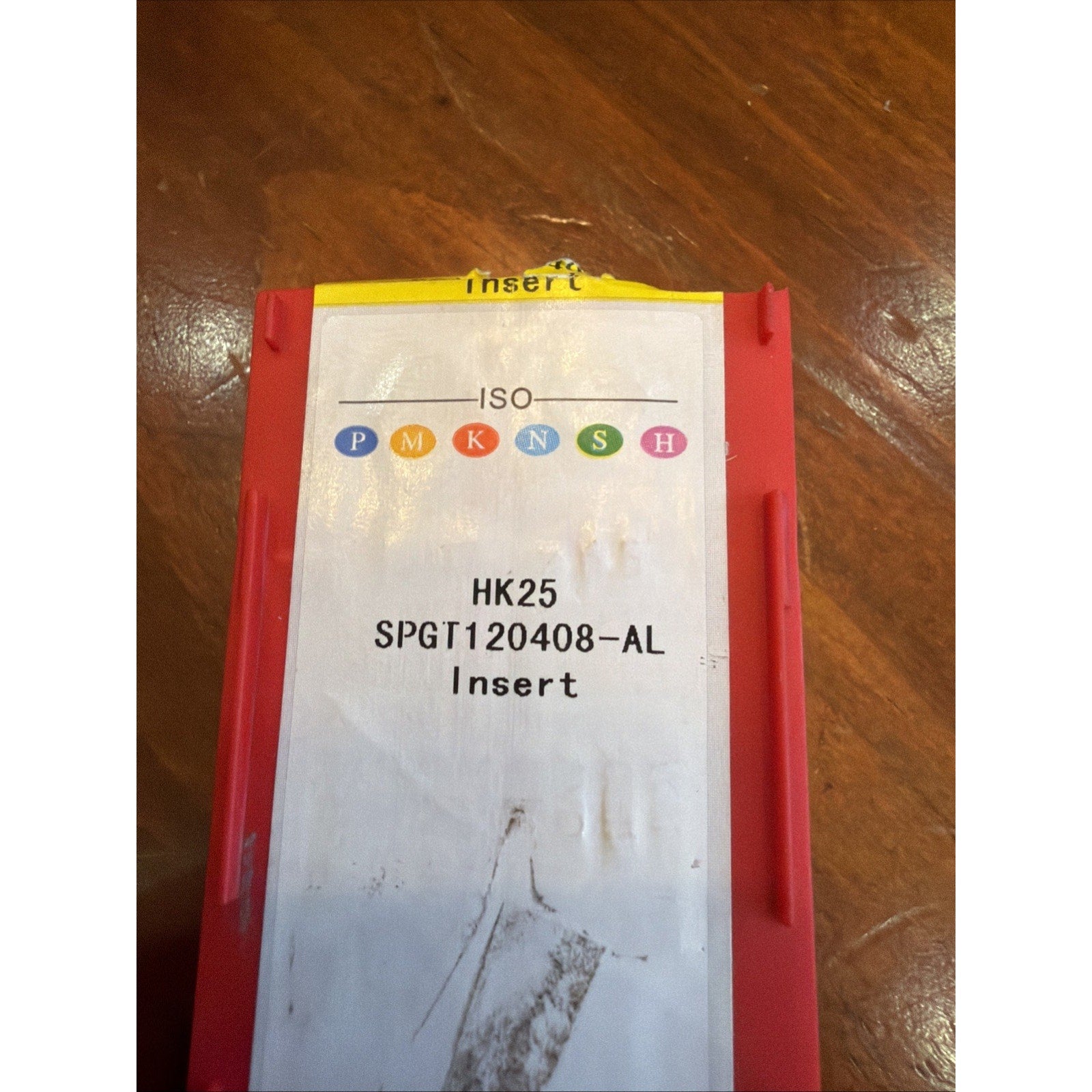 Red plastic storage container with a white label showing ISO color-coded classifications and text "HK25 SPGT120408-AL Insert," placed on a wooden surface.