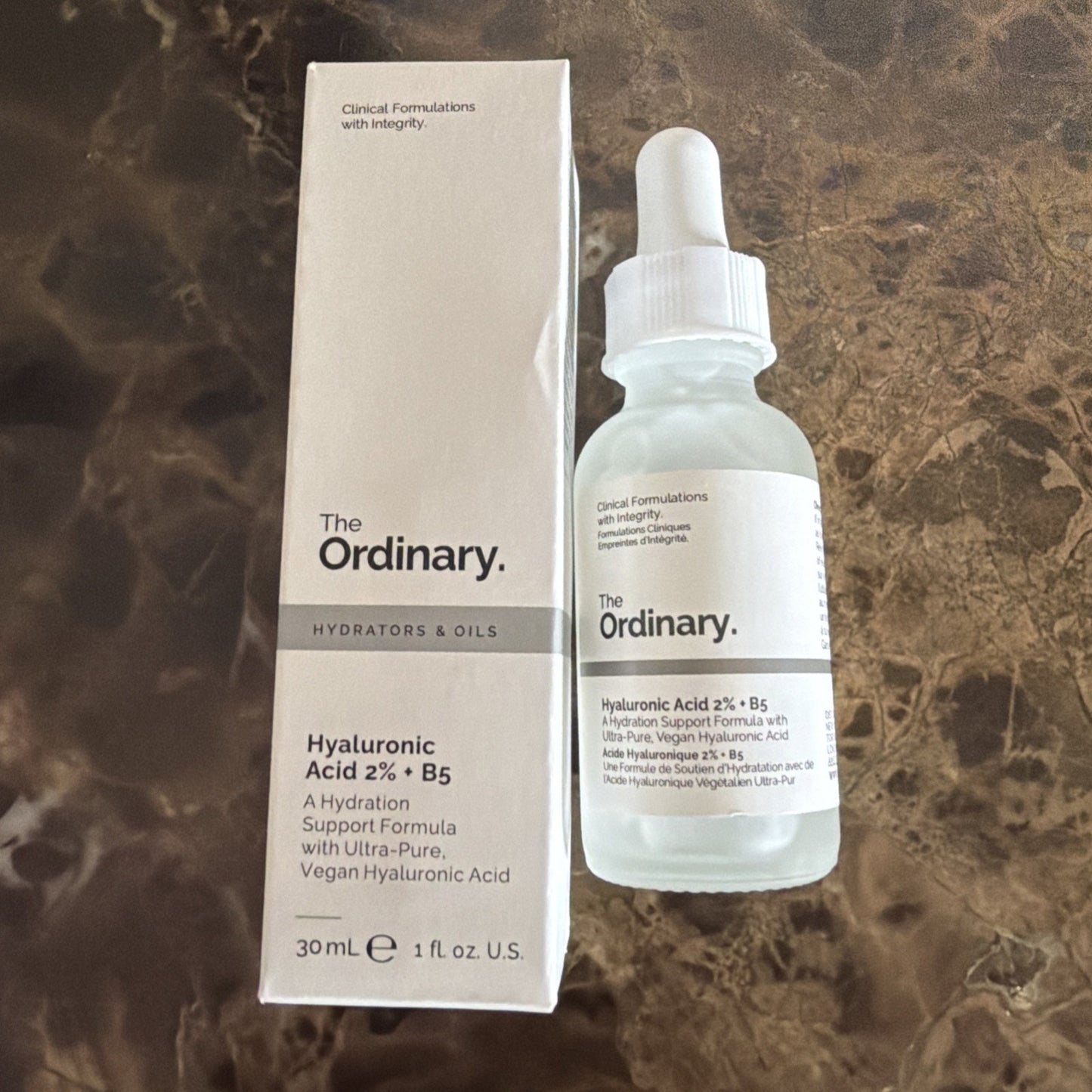 Glass dropper bottle and white rectangular box of The Ordinary Hyaluronic Acid 2% + B5 serum, featuring minimal black text and placed on a brown marbled surface.