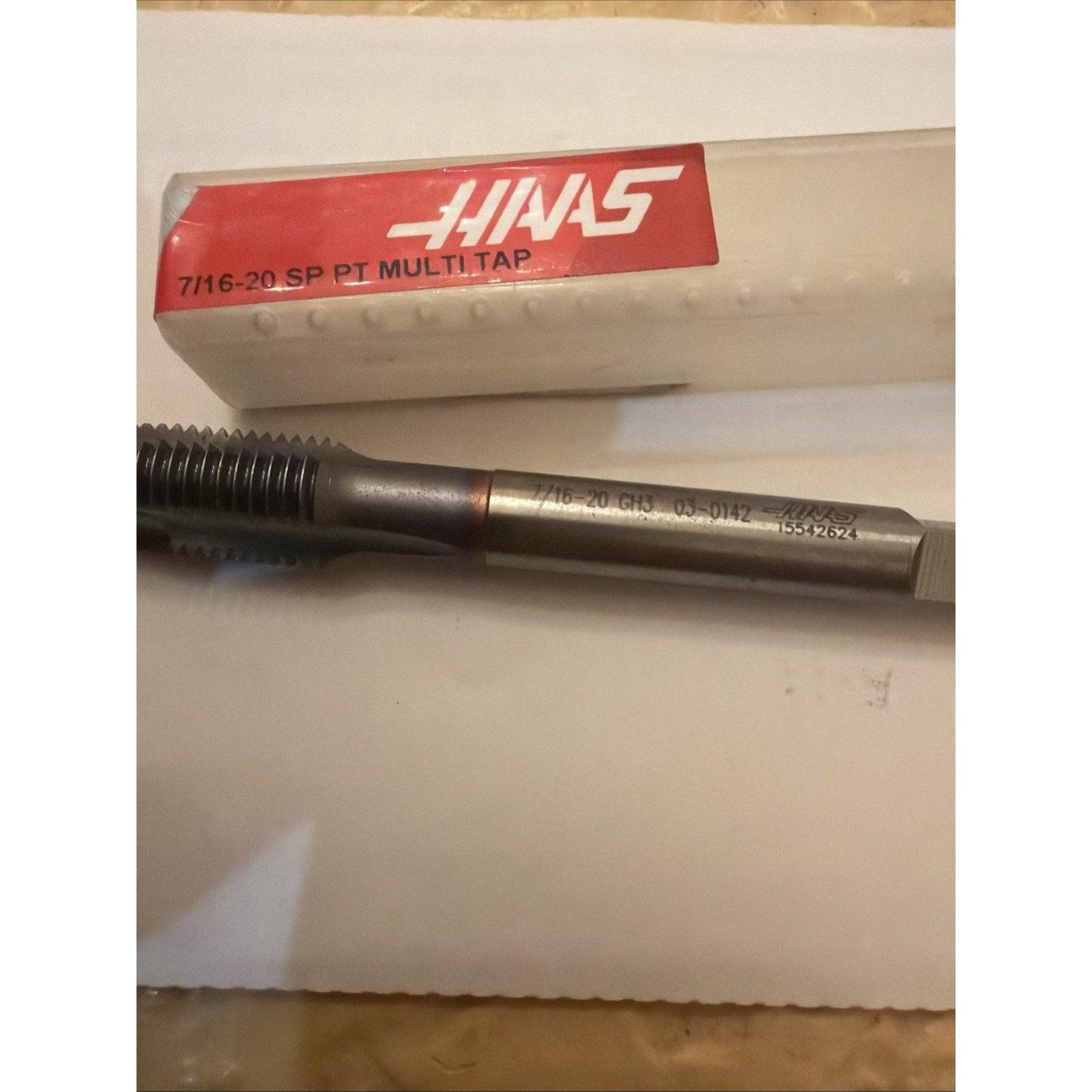 Metal threading tap with sharp spiral threads and a cylindrical shank featuring engraved size and product numbers, placed near a white plastic case with a red Haas label.