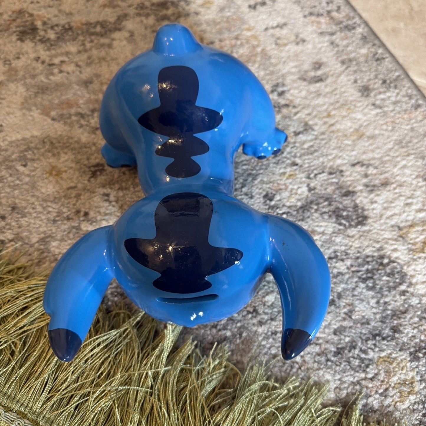 Glossy blue ceramic figure with black top hat silhouettes painted on the back and outstretched arms, resting on a textured carpet near a green tassel accessory.