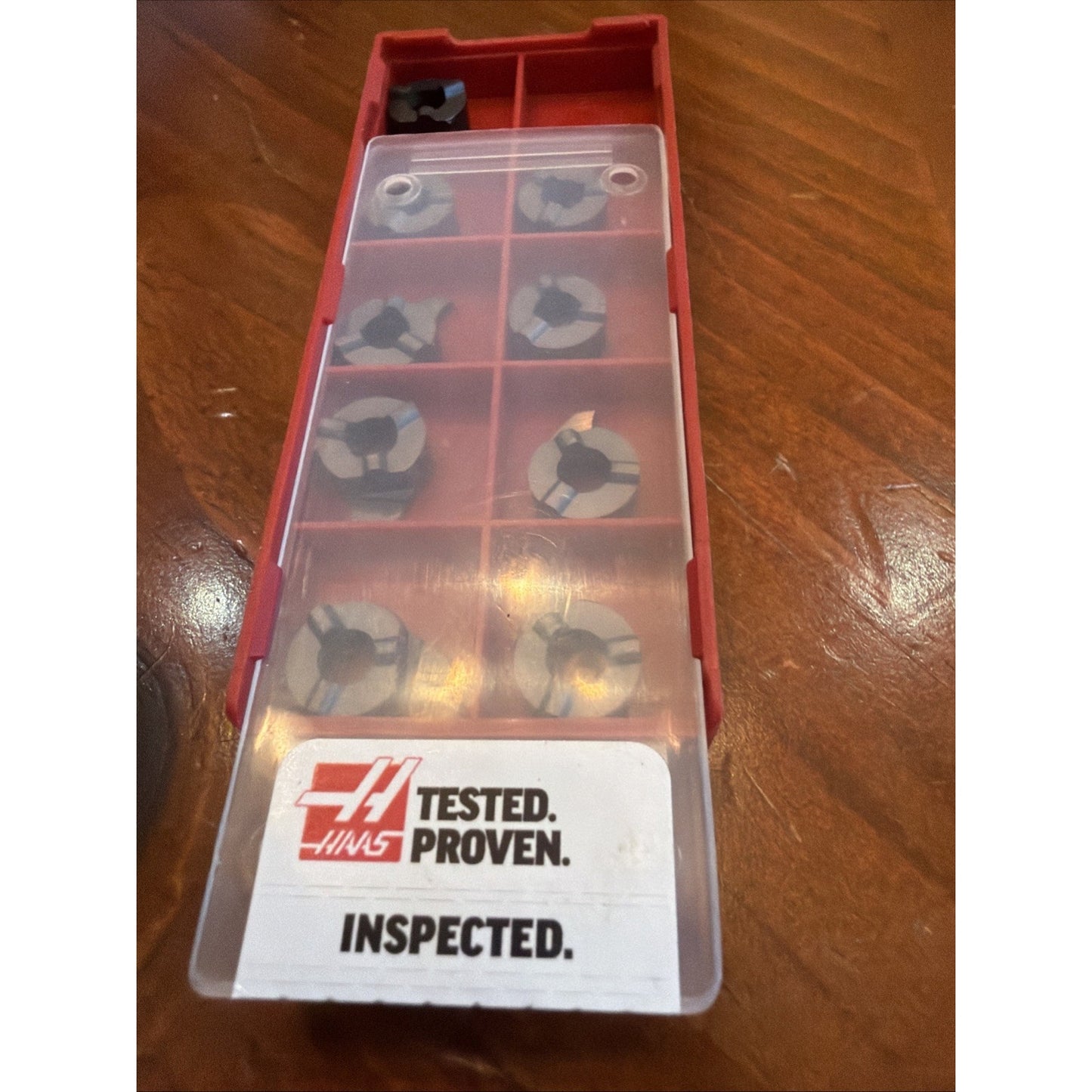 Red plastic storage box with a translucent lid containing eight round segmented metal inserts arranged in separate compartments, featuring a Haas Tested Proven Inspected label.