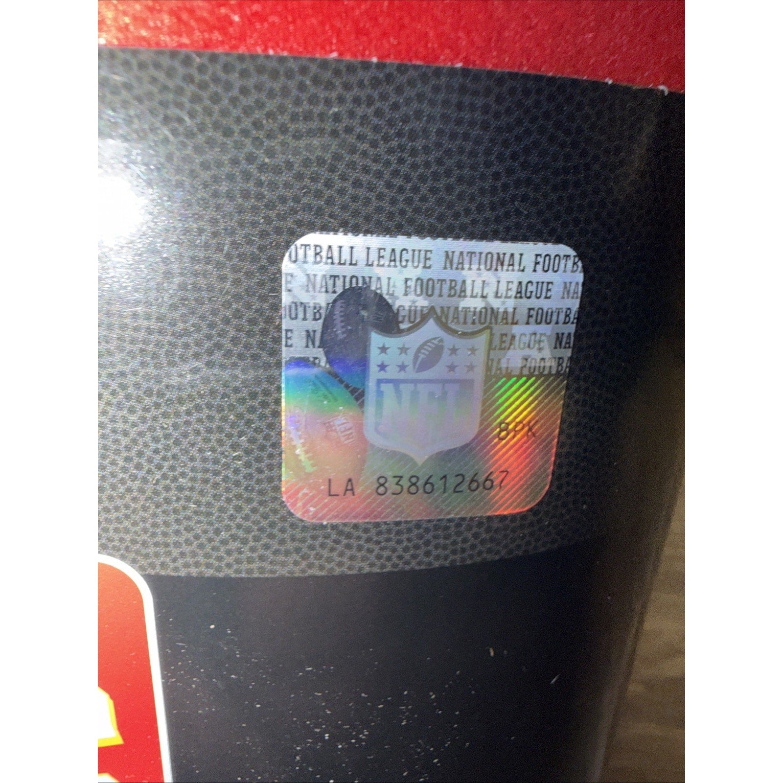 Close-up of a holographic NFL authenticity sticker with the NFL shield logo and serial number on black textured packaging resembling a football surface.