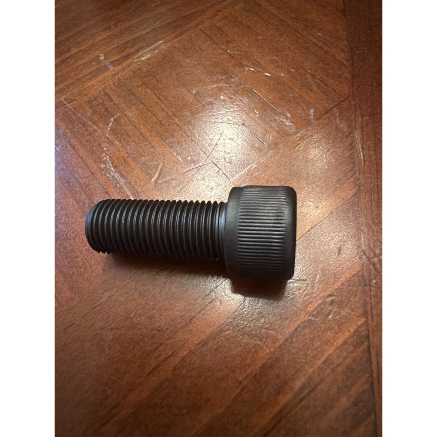 Black threaded screw with a large knurled head resting on a wooden surface with a visible grain pattern.
