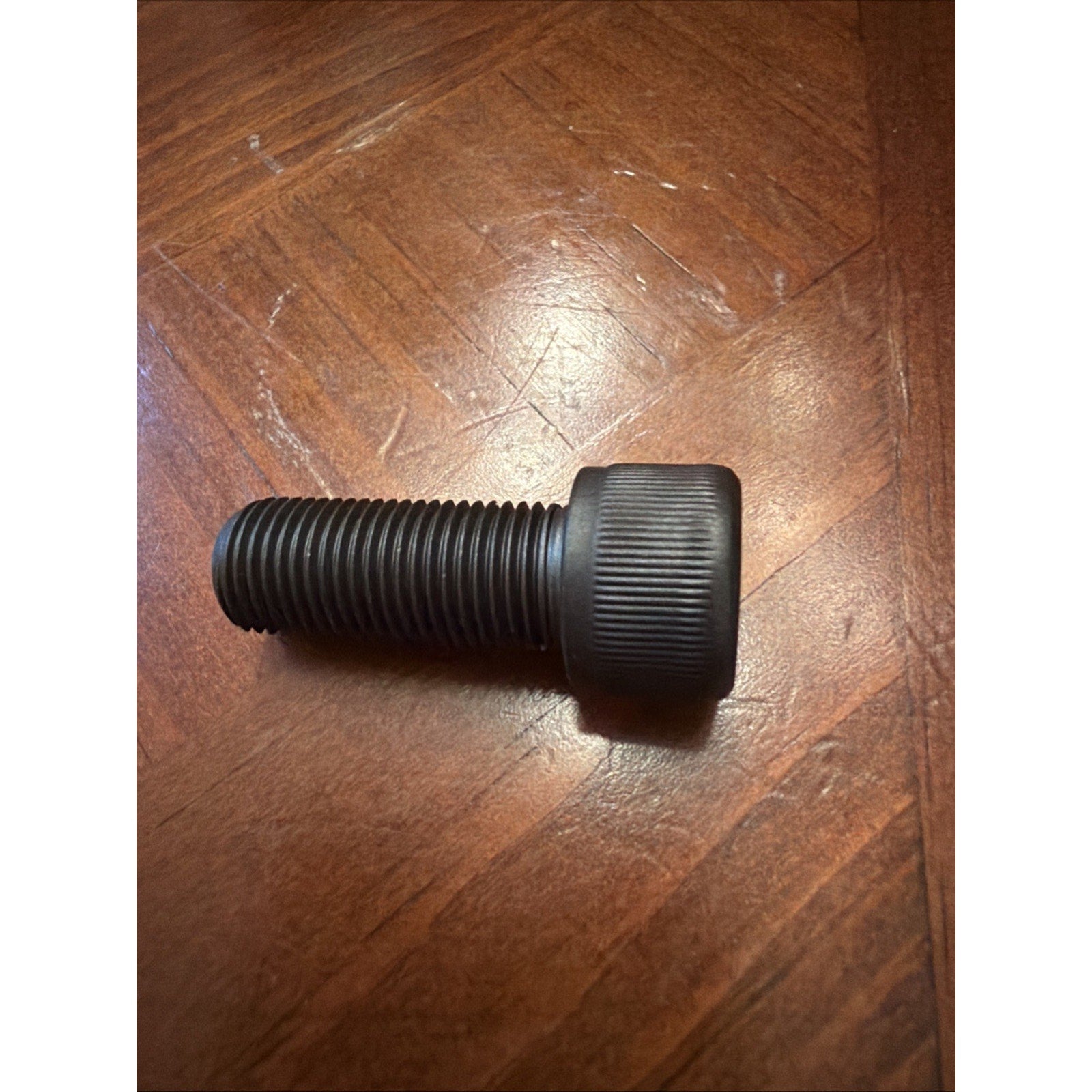 Black threaded screw with a large knurled head resting on a wooden surface with a visible grain pattern.
