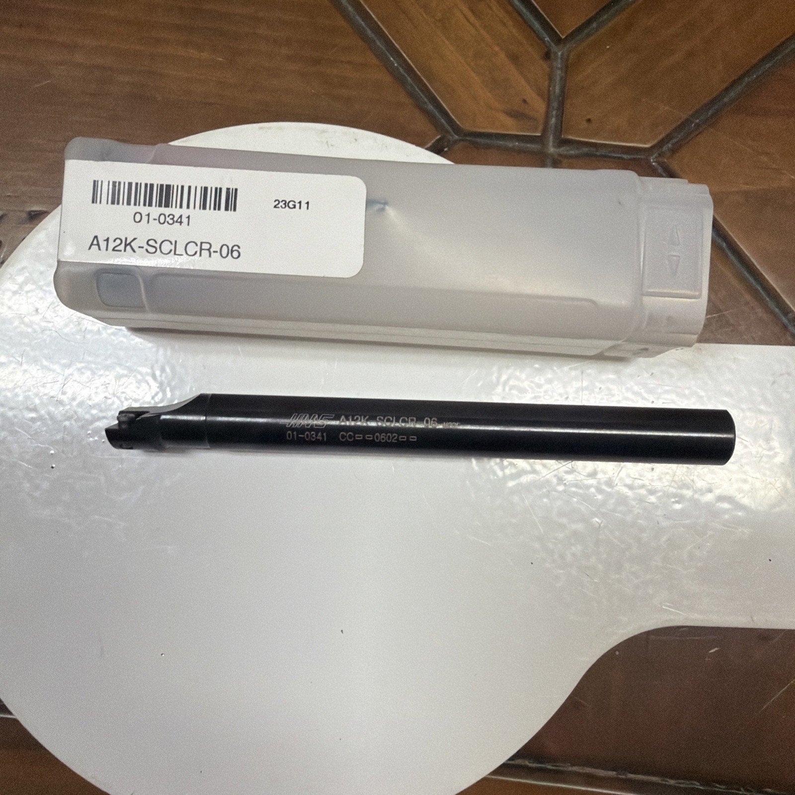 Black metal cutting tool engraved with the code "A12K-SCLCR-06," placed on a white surface next to a frosted plastic storage case with a barcode label.