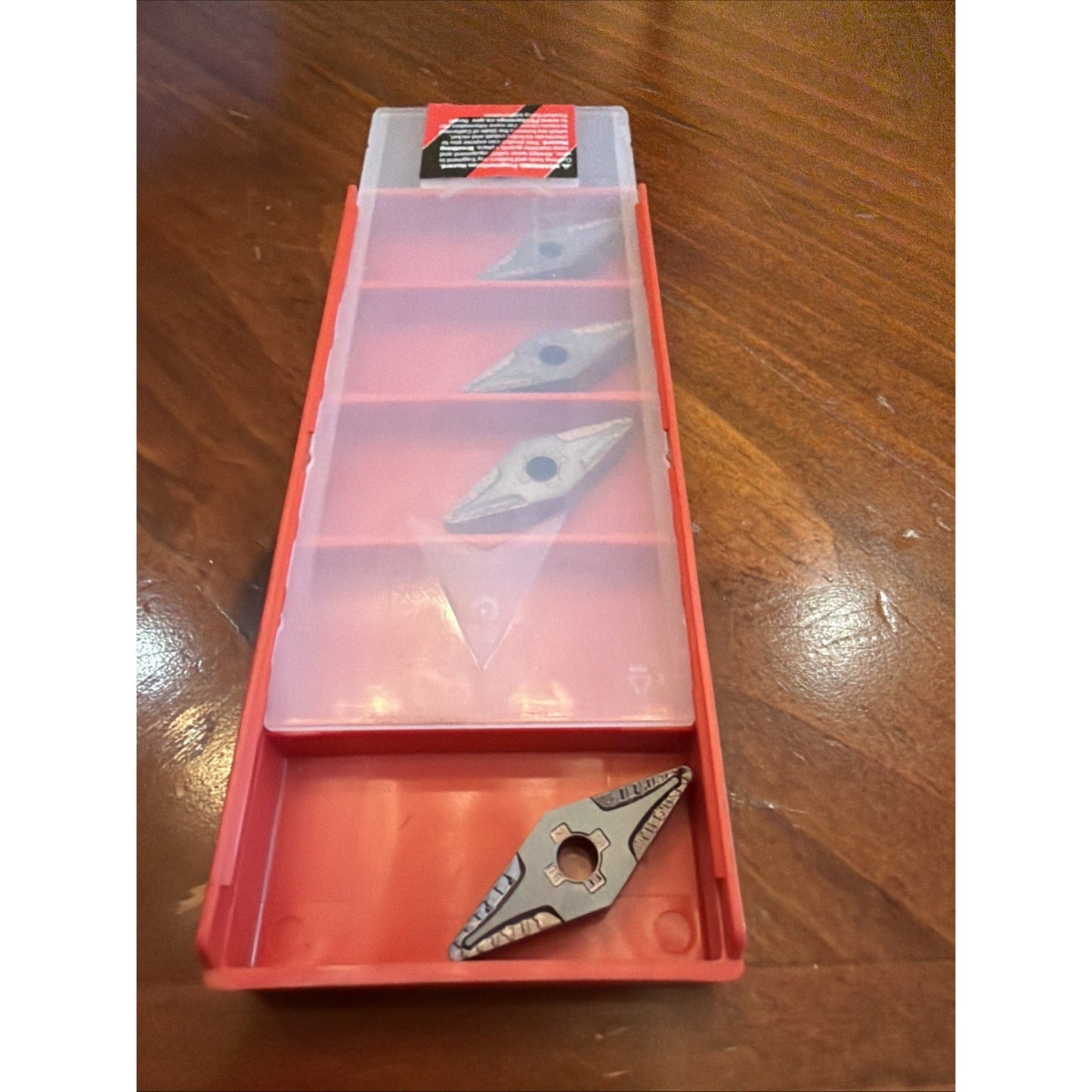 Long red plastic container with a translucent lid holding four uniquely shaped metal cutting inserts arranged in separate compartments on a wooden surface.