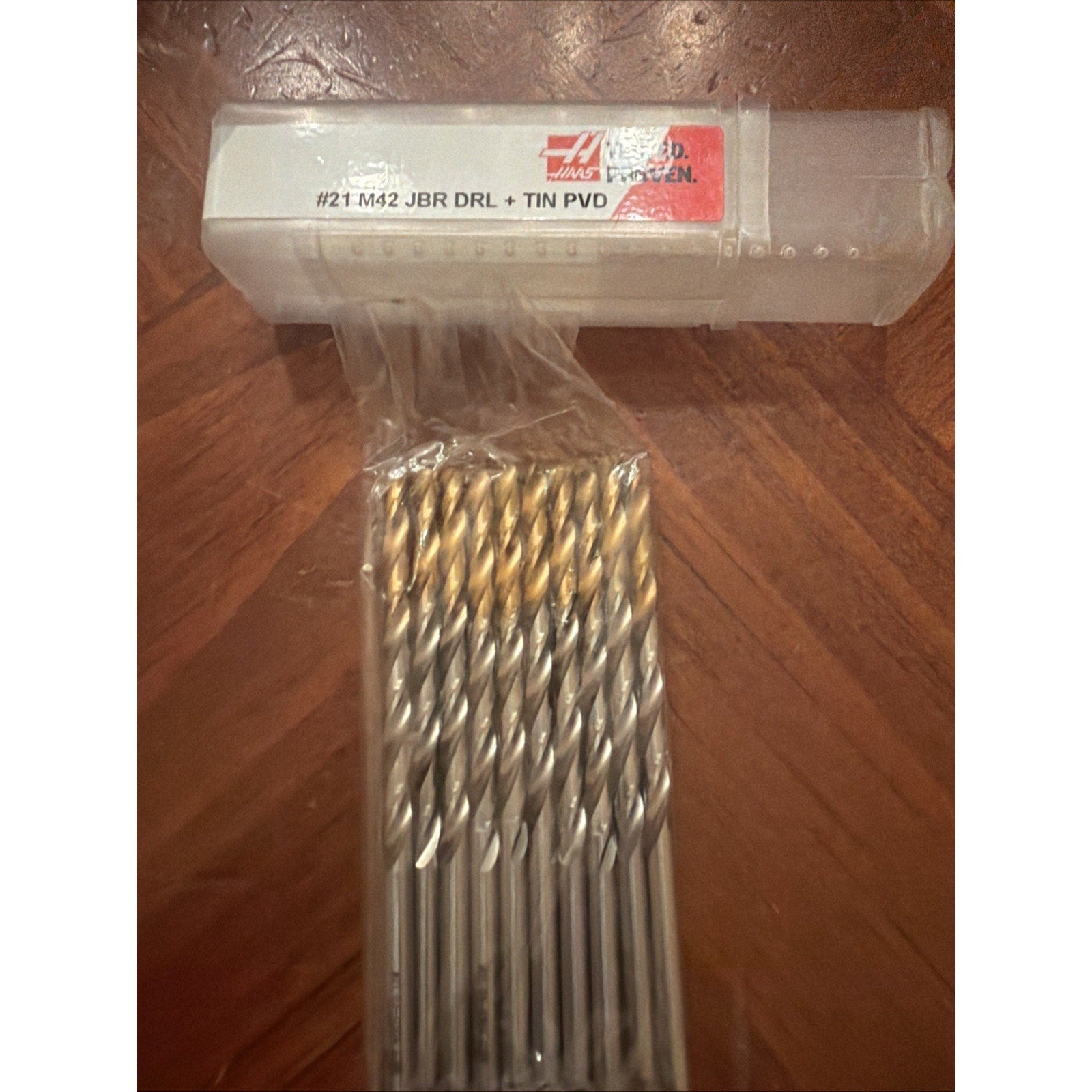 Set of multiple metal drill bits with gold-colored coated tips arranged inside a clear plastic case labeled with product specifications, placed on a wooden surface.