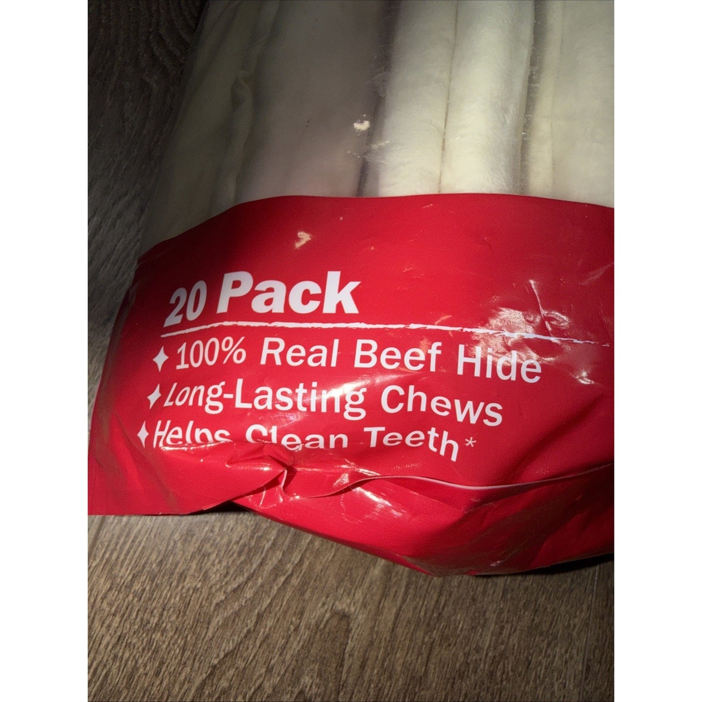 Partial view of a pack of white rawhide dog chews wrapped in clear plastic with a red label stating "20 Pack," "100% Real Beef Hide," and "Long-Lasting Chews" on a wooden surface.