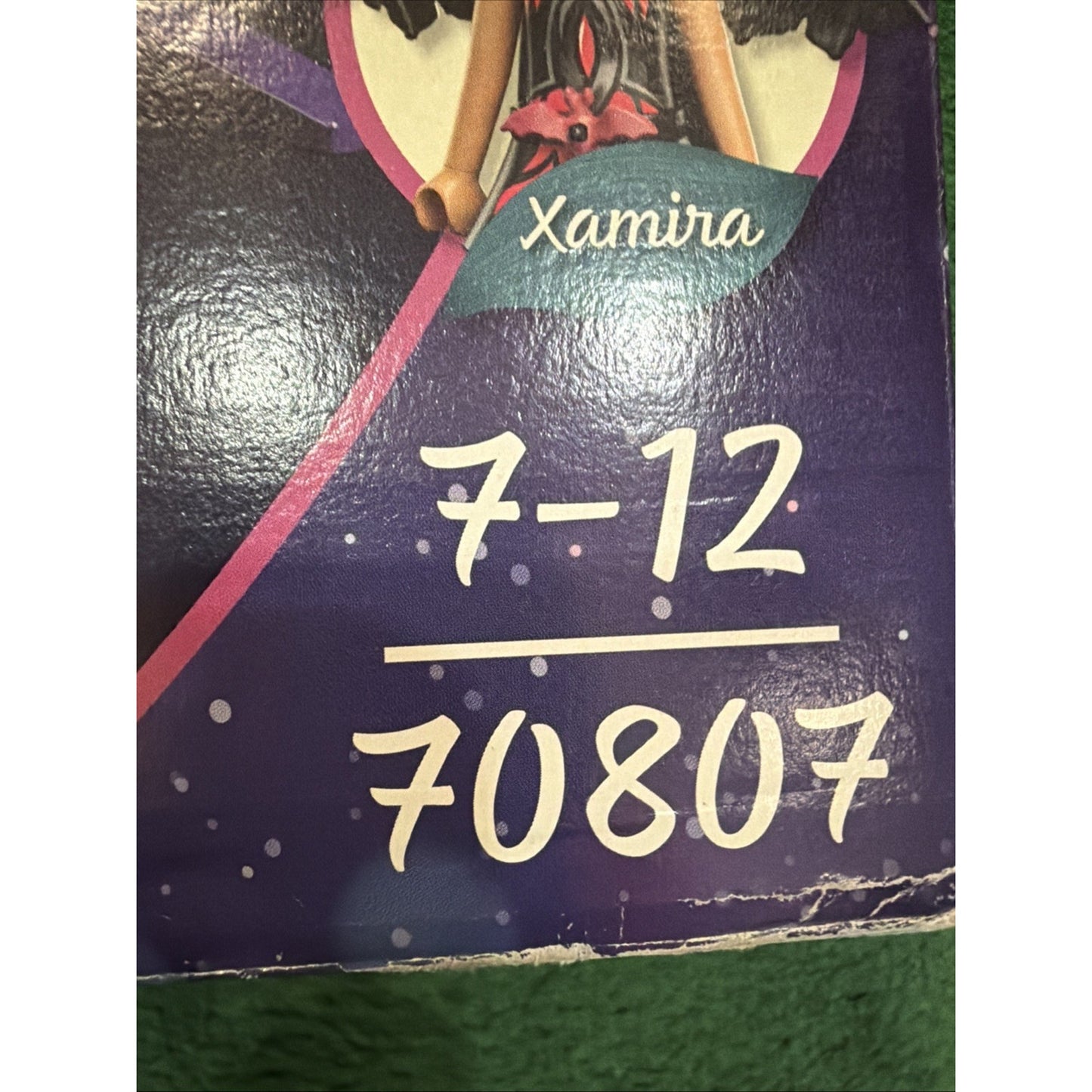 Close-up of a purple box corner showing the name "Xamira," age range "7-12," and product number "70807" in white handwritten-style text on a textured surface.