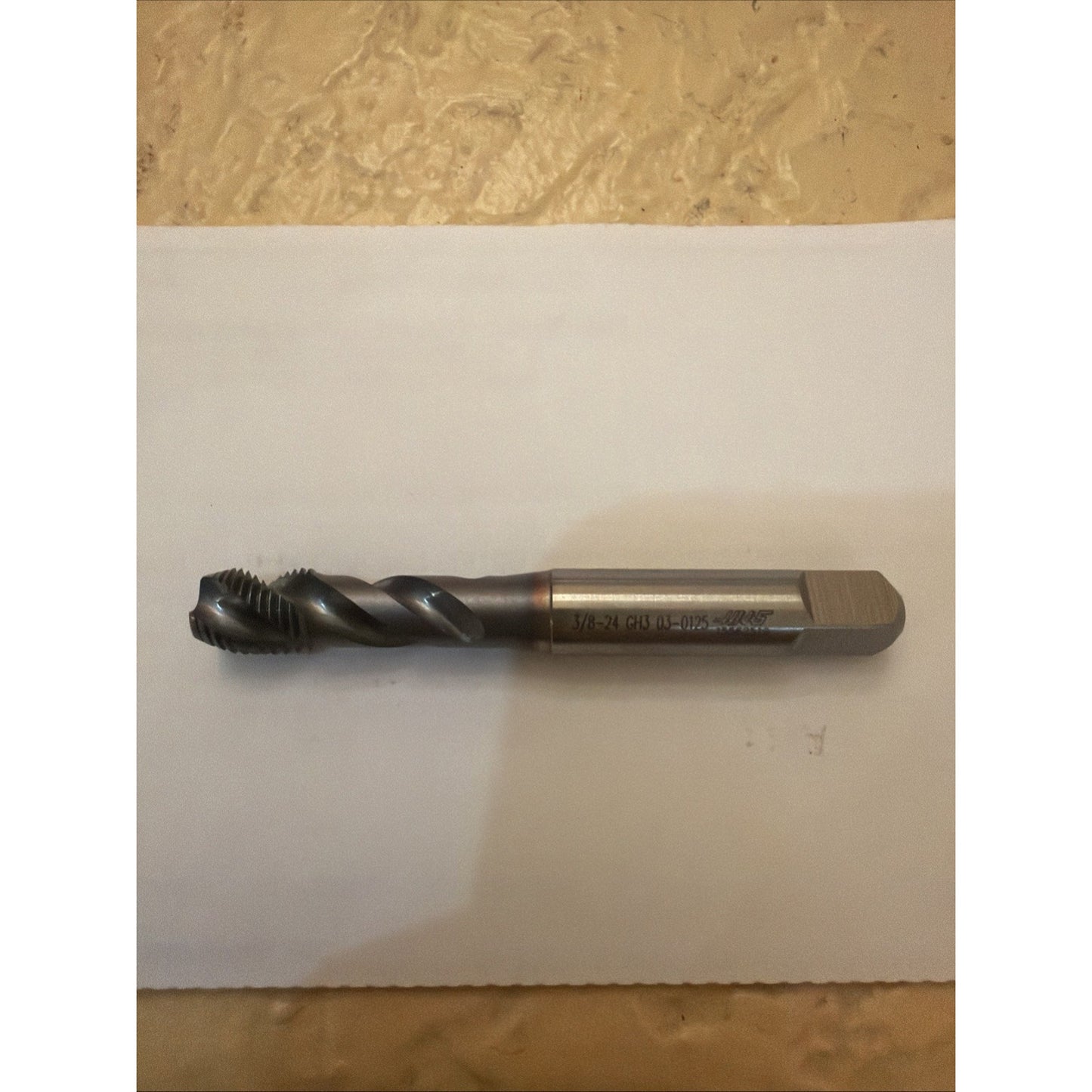 Single metal spiral threading tap with a square shank and sharp cutting threads, resting horizontally on a white sheet against a textured beige background.