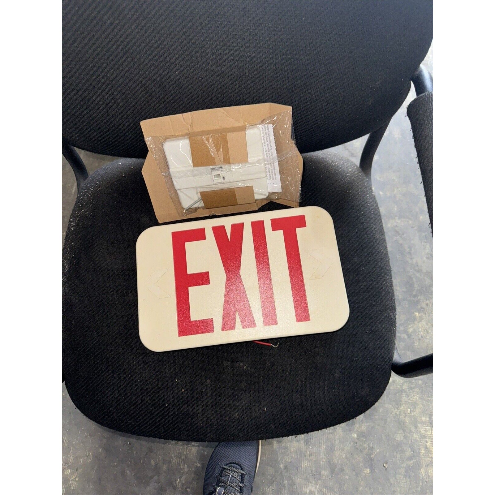 Commercial Electric LED exit sign with red EXIT letters and cardboard packaging on black fabric chair seat