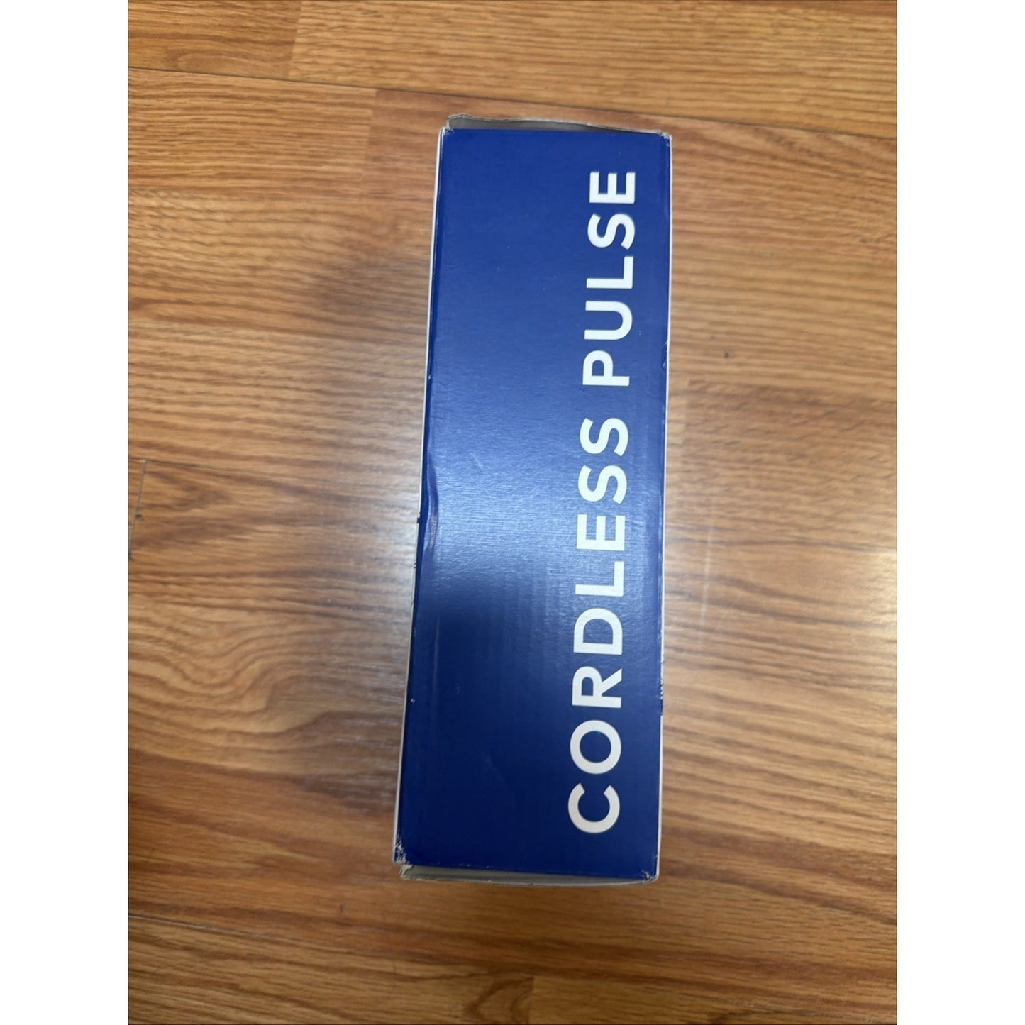 Blue rectangular box with the words "Cordless Pulse" printed in white capital letters on a wooden floor background.