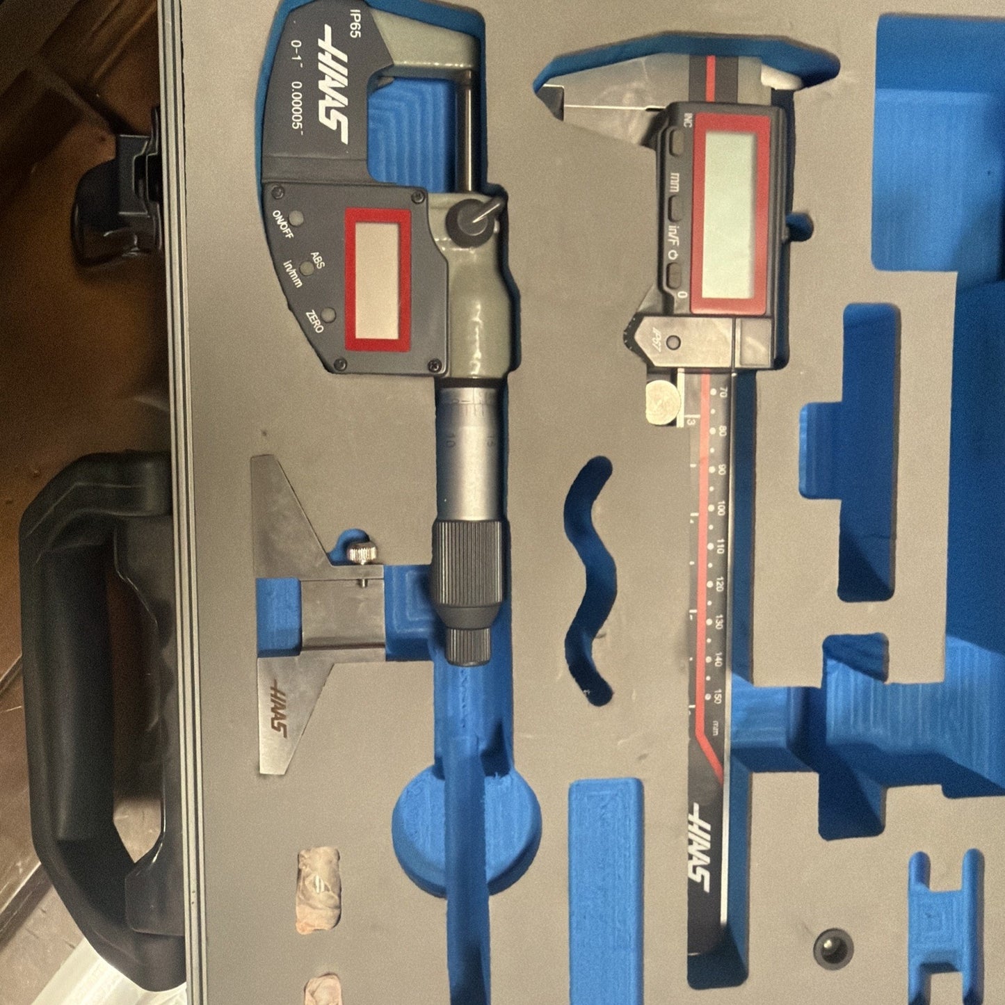 Open gray carrying case with blue custom foam insert securely holding two digital measuring tools, a metal triangular calibration gauge, and two small cylindrical objects on a wooden surface.