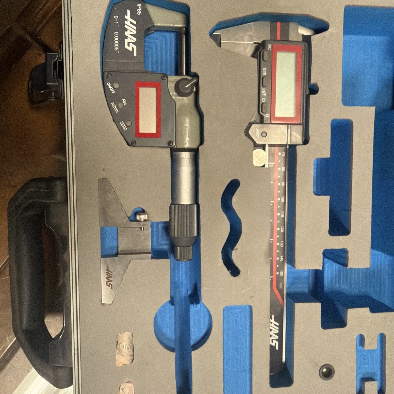 Open gray carrying case with blue custom foam insert securely holding two digital measuring tools, a metal triangular calibration gauge, and two small cylindrical objects on a wooden surface.