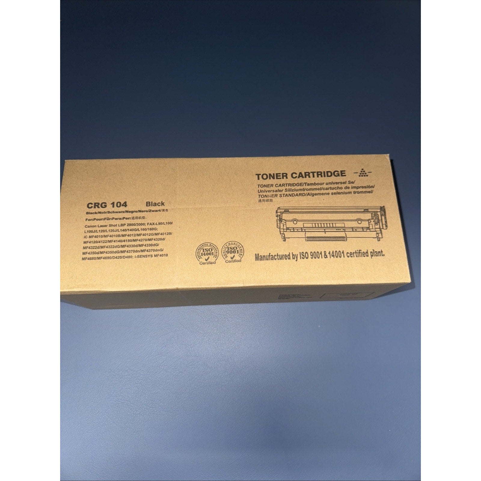 Brown cardboard box packaging for a black toner cartridge labeled CRG 104 with product details, certification logos, and an illustration of the cartridge on a blue surface background